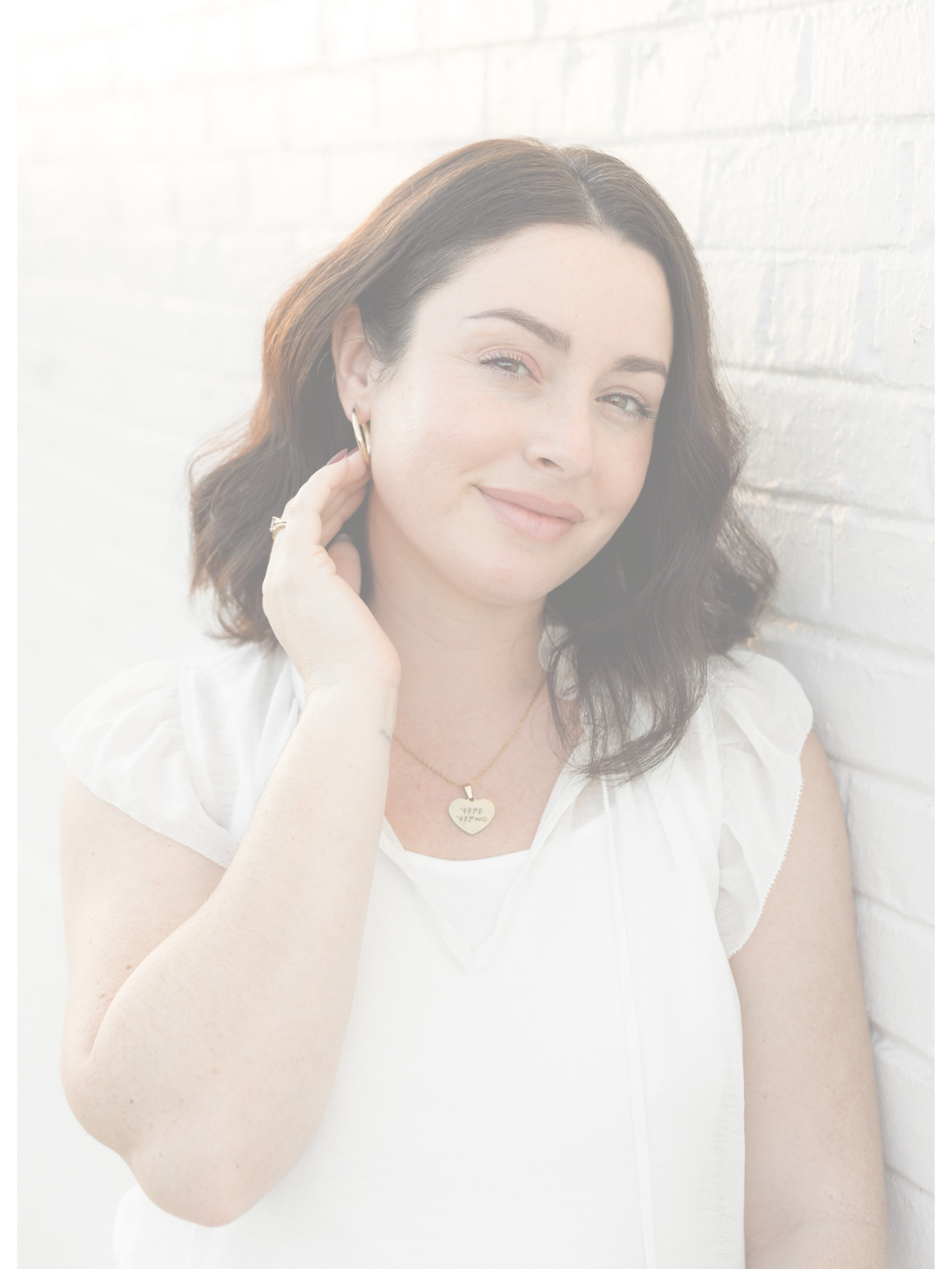 Woman with dark brown hair wearing white top and gold jewelry, leaning against a white brick wall, smiling softly.