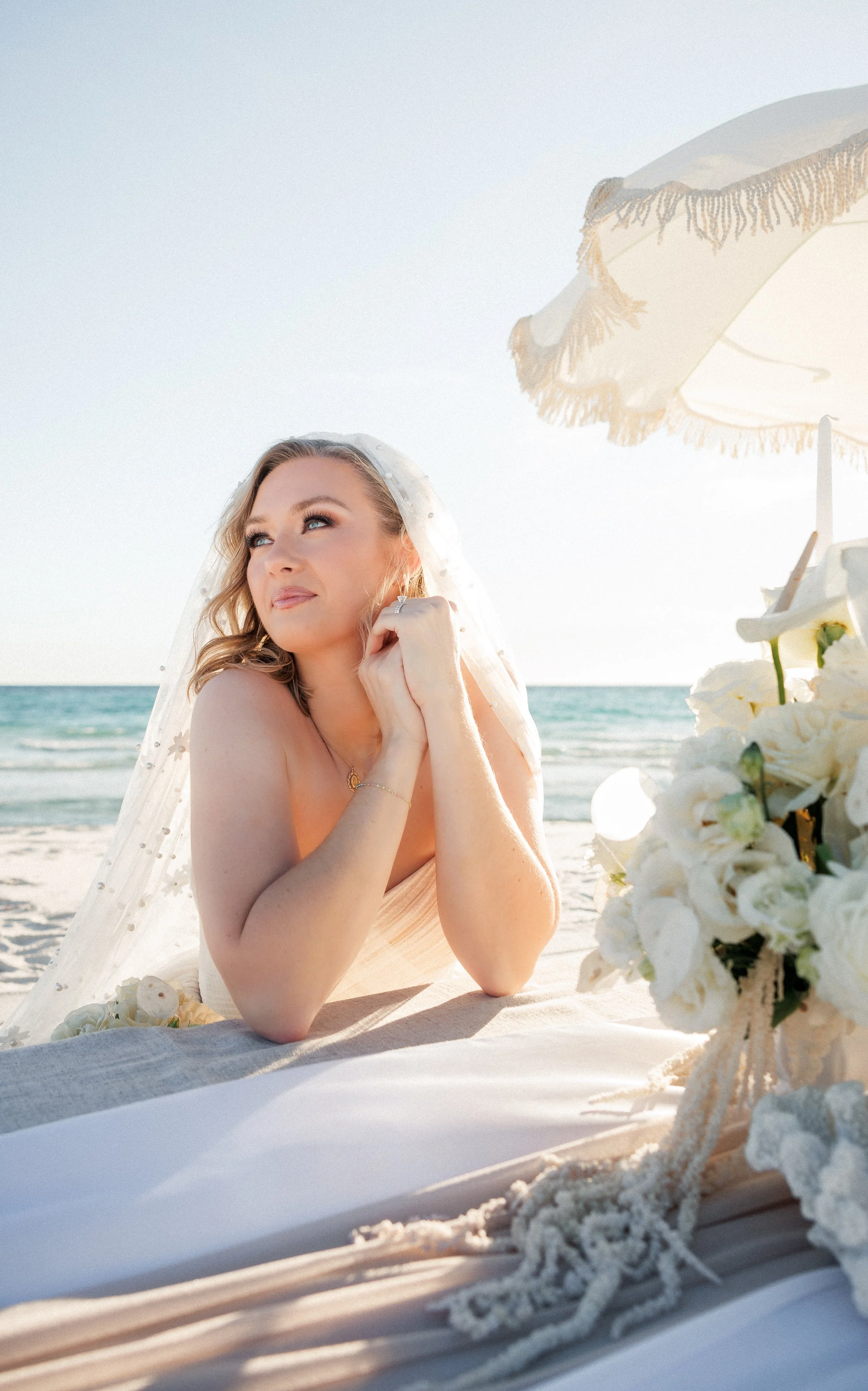 A woman in a wedding dress with a veil, sitting on the beach near the ocean, under a white umbrella, with a floral arrangement nearby.