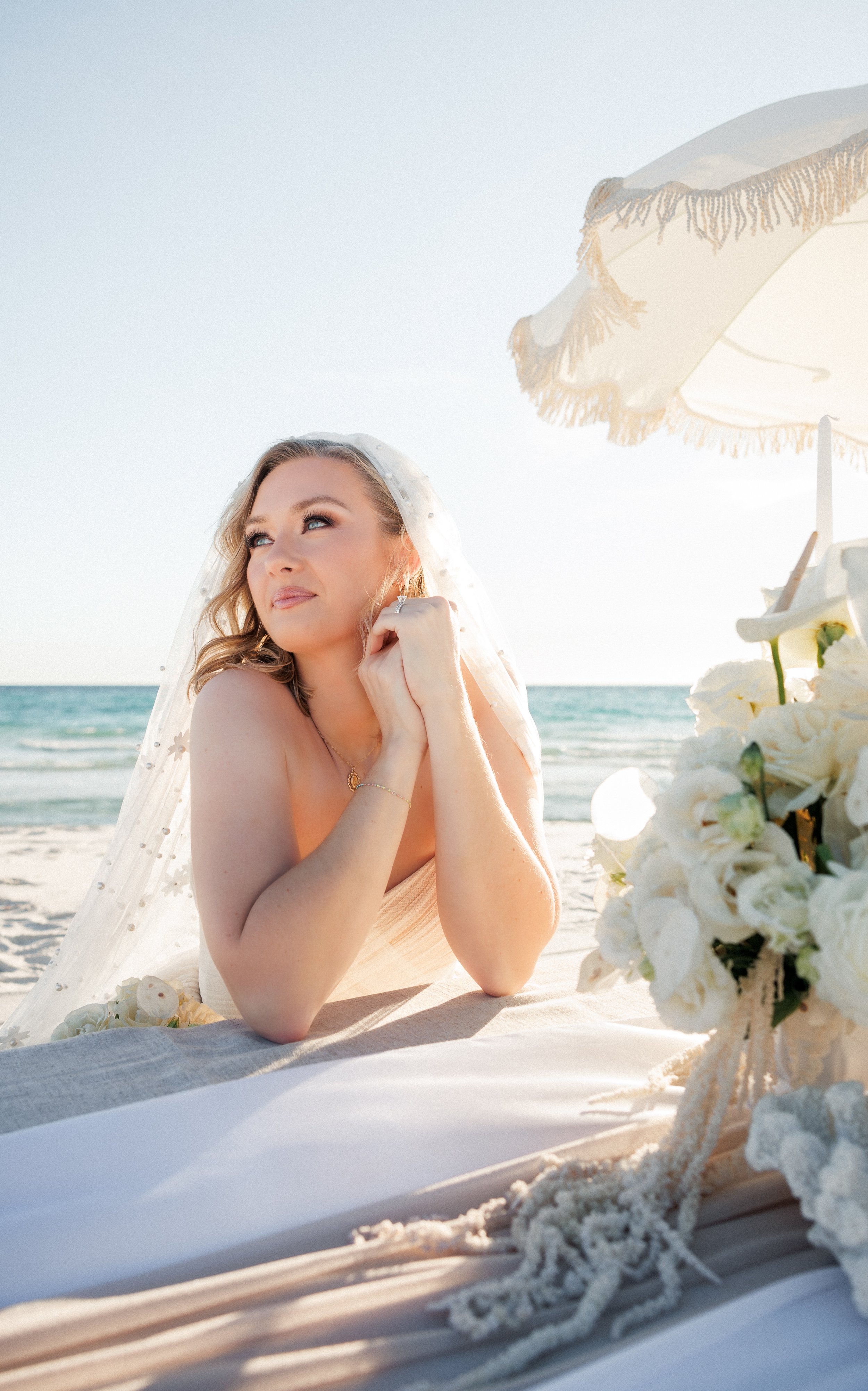 A woman in a wedding dress with a veil, sitting on the beach near the ocean, under a white umbrella, with a floral arrangement nearby.