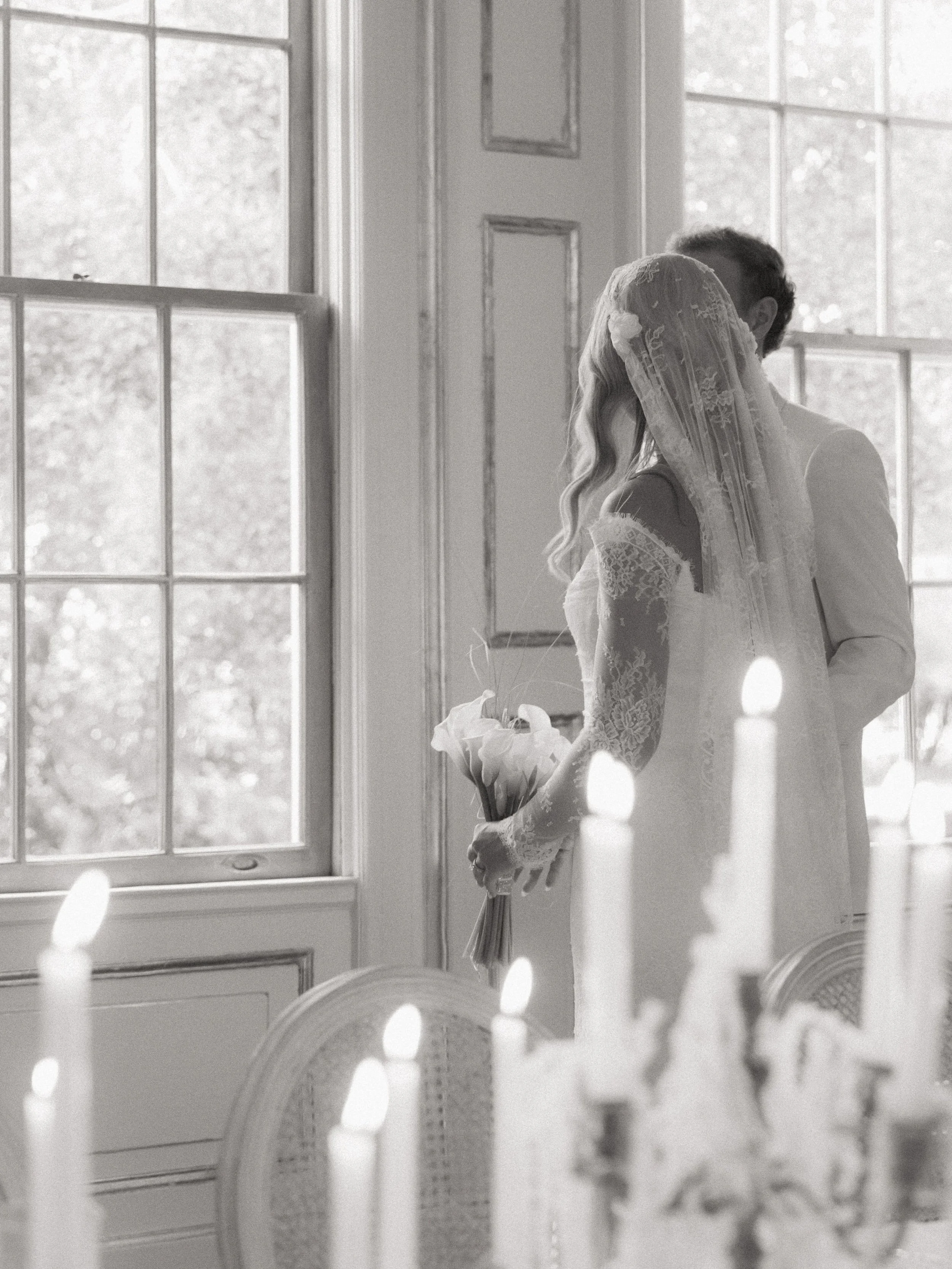 A bride and groom share a kiss in a warmly lit room with large windows, a bride holding a bouquet of flowers, and candles in the foreground.