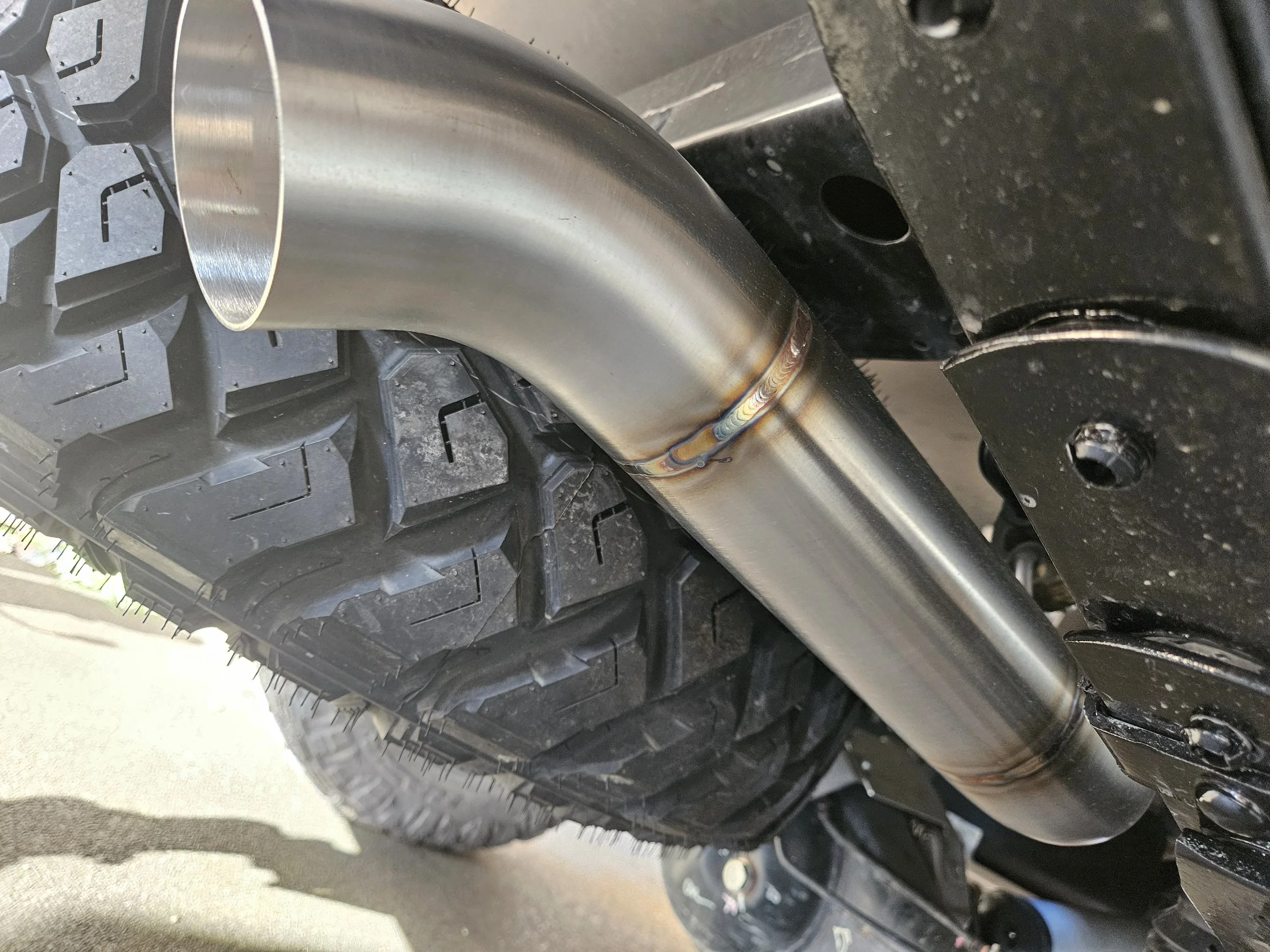 Close-up of a metallic exhaust pipe mounted under a vehicle, positioned near a rugged off-road tire.