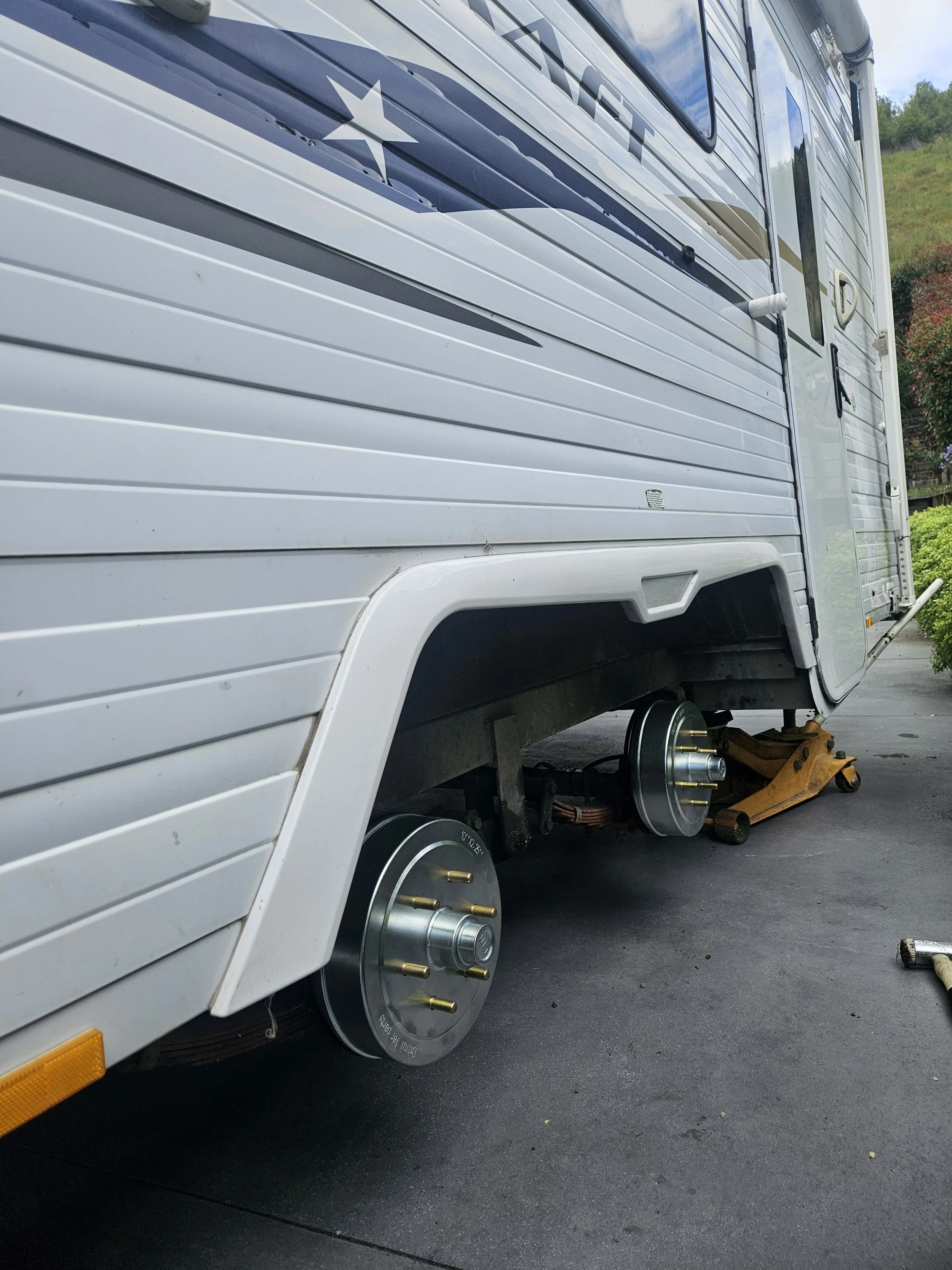 The side of a white RV with wheels removed, showing brake drums and a jack lifting the vehicle.