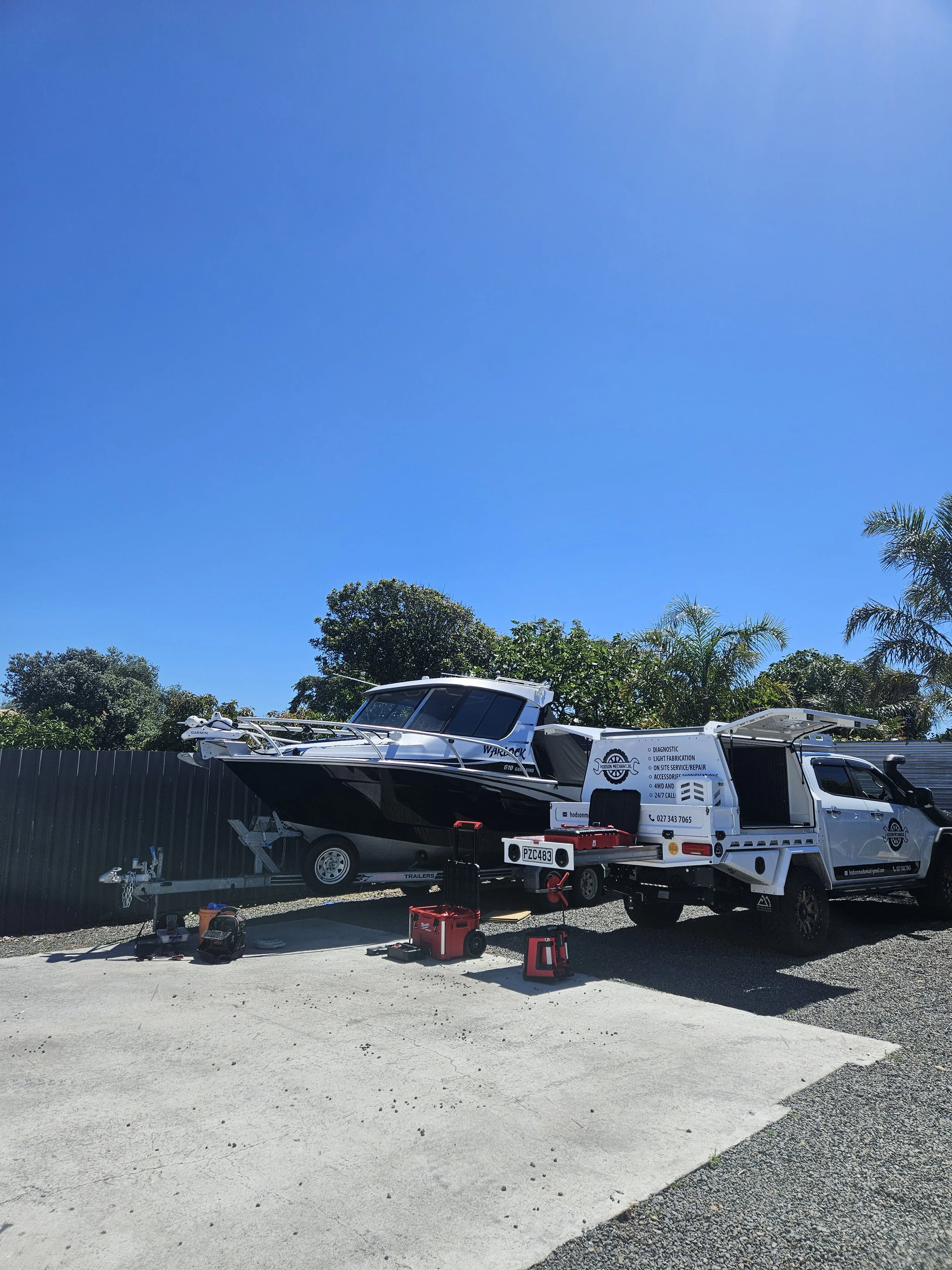 A boat on a trailer hitched to a white pickup truck, parked outdoors under a clear blue sky with trees in the background.