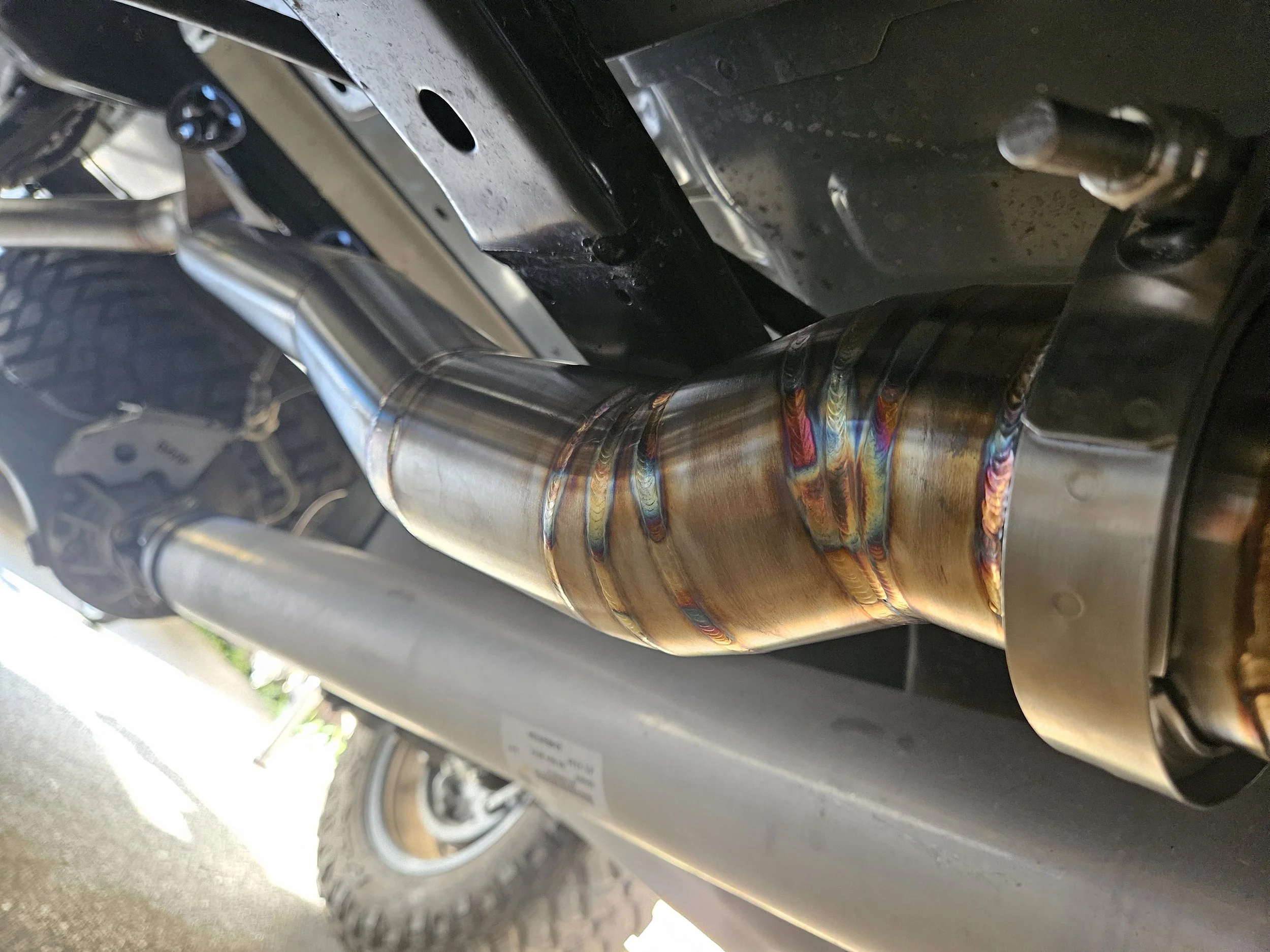 Close-up view of a metal exhaust pipe with colorful heat discoloration on a vehicle, with a part of the vehicle's tire and frame visible in the background.