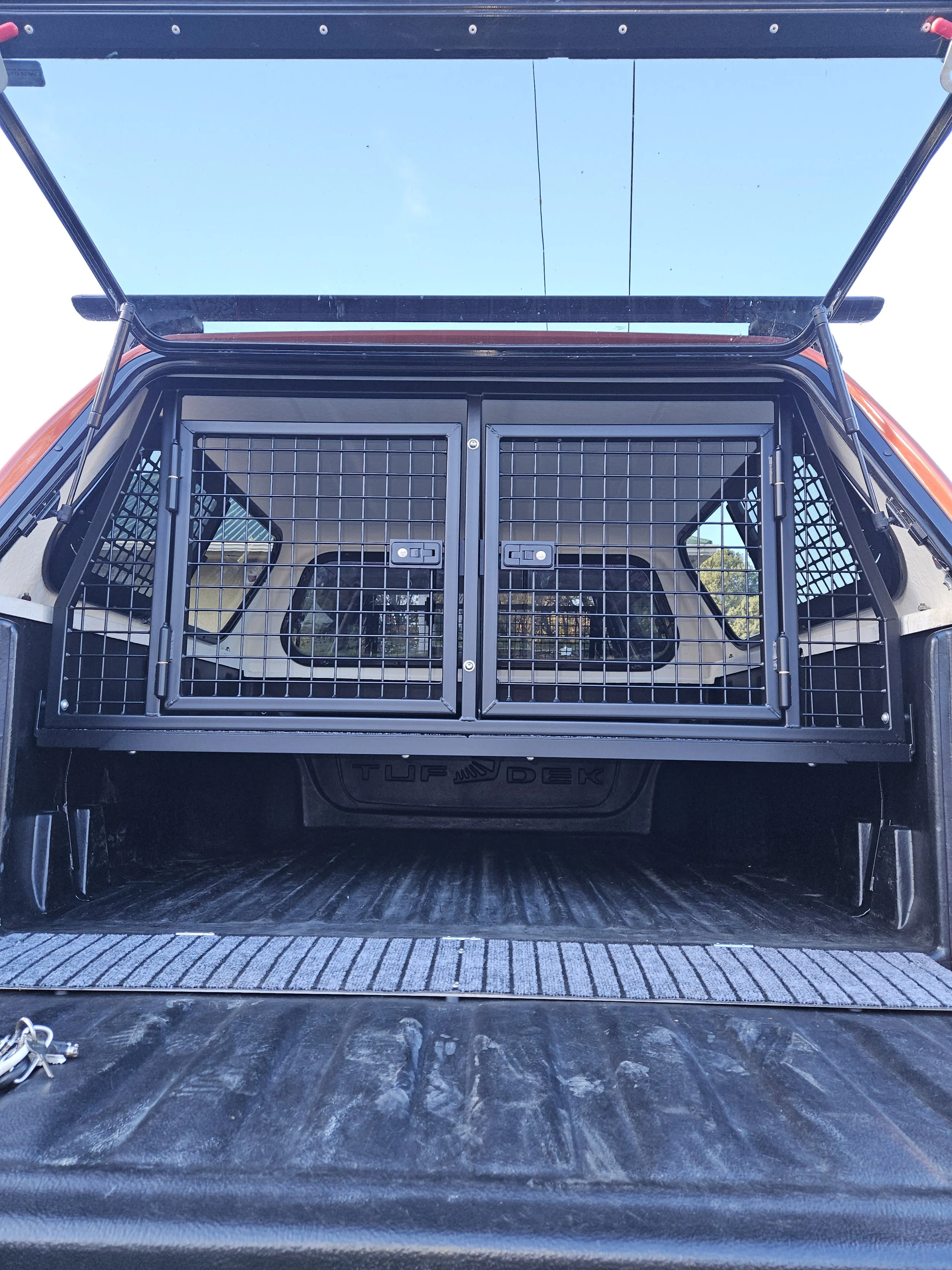 Back of a pickup truck with a metal pet crate secured in the truck bed, and the tailgate open under a blue sky.