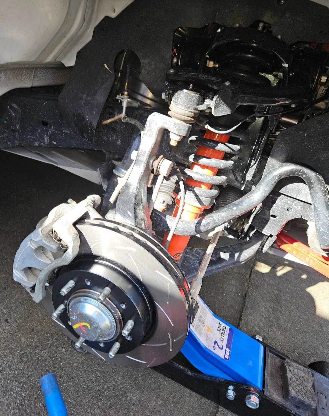 A vehicle's front wheel has been removed, showing the brake disc, caliper, suspension, and strut assembly.