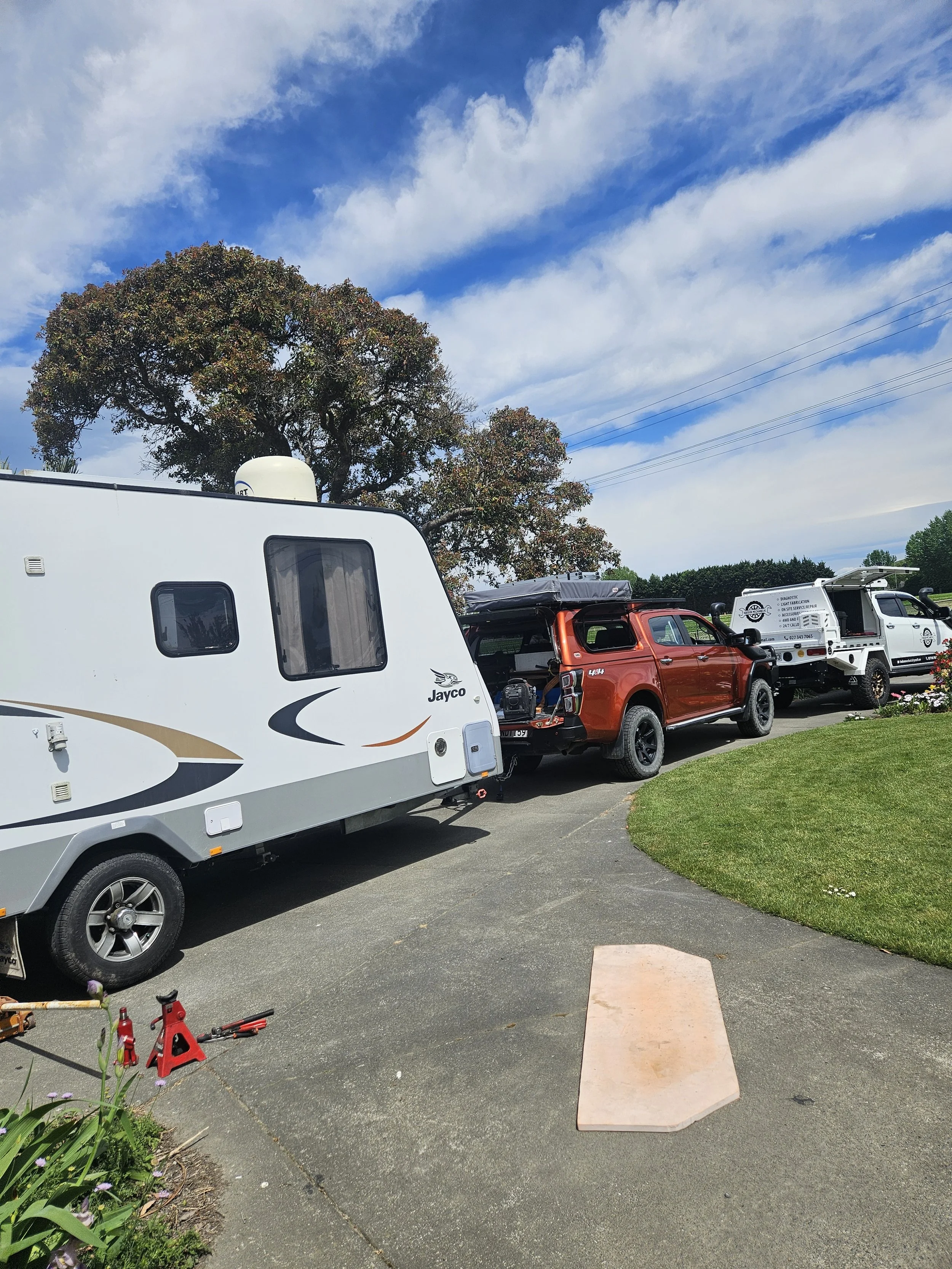 A campground driveway with a white travel trailer, a red pickup truck, and a white utility vehicle parked on a curving asphalt road next to a grassy lawn and a large tree under a partly cloudy sky.