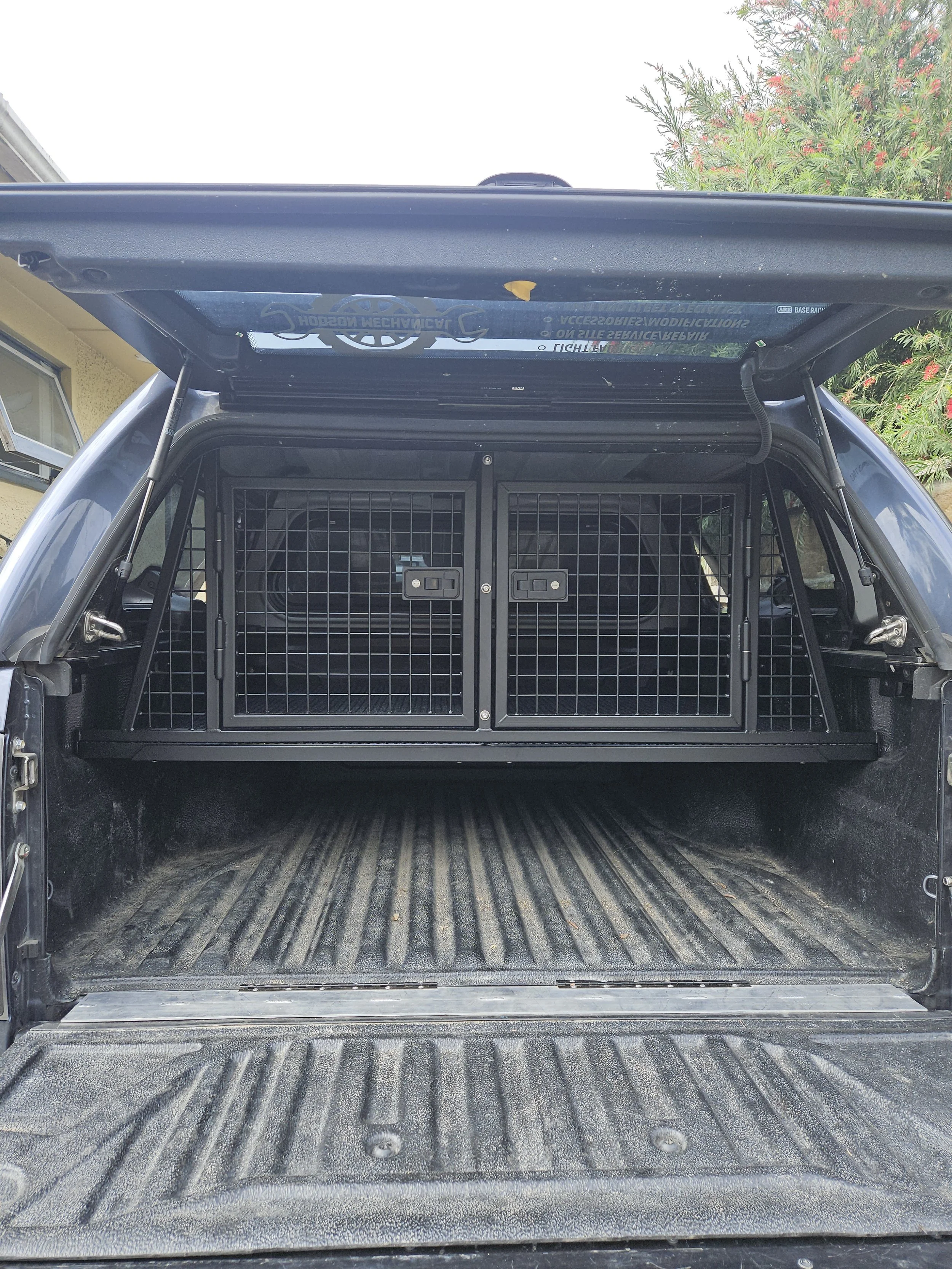 The open tailgate of a vehicle shows a pet crate in the back, with a metal mesh door.