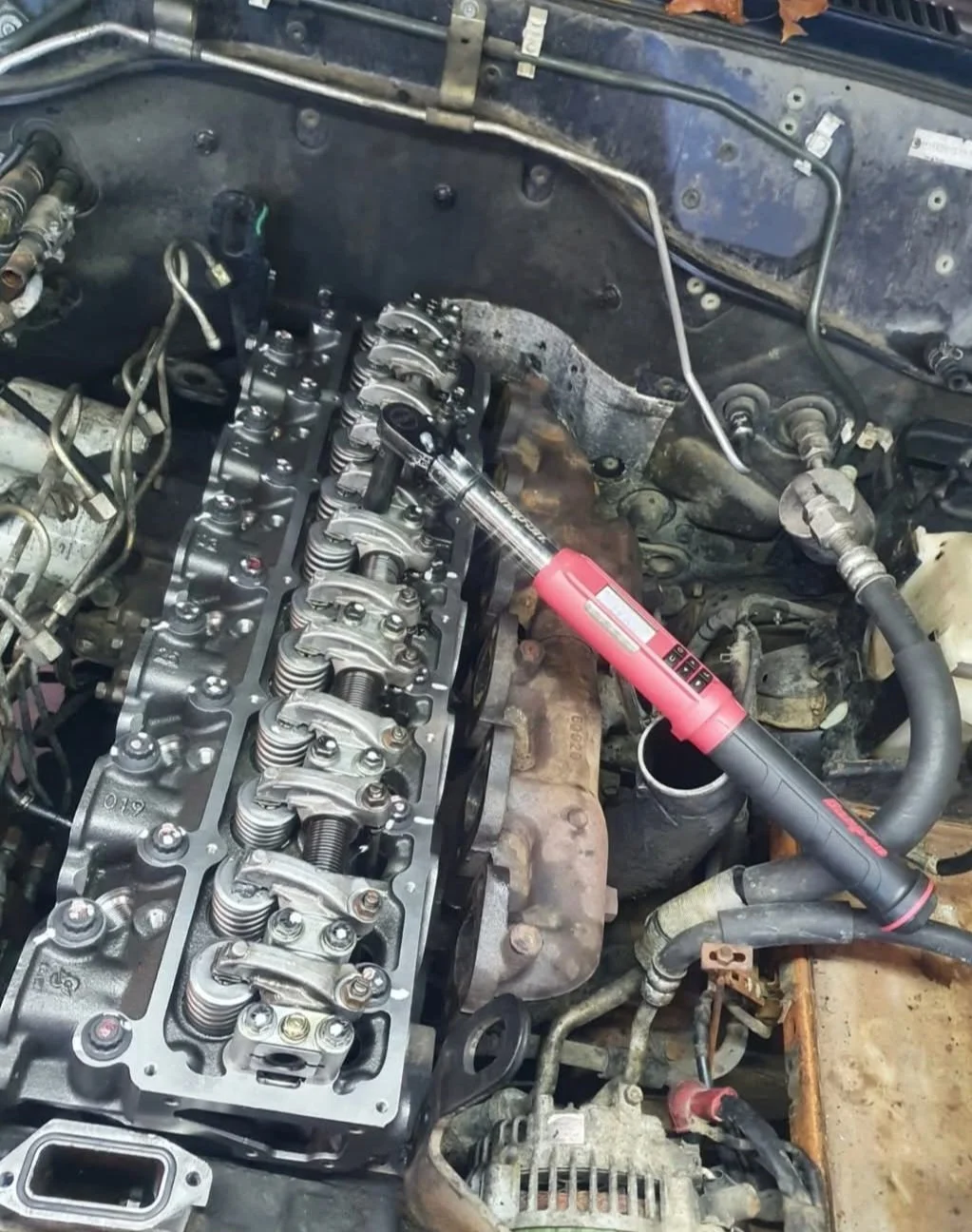 A car engine with a cylinder head removed, showing valves and valve springs, with a red impact wrench resting on the engine block.