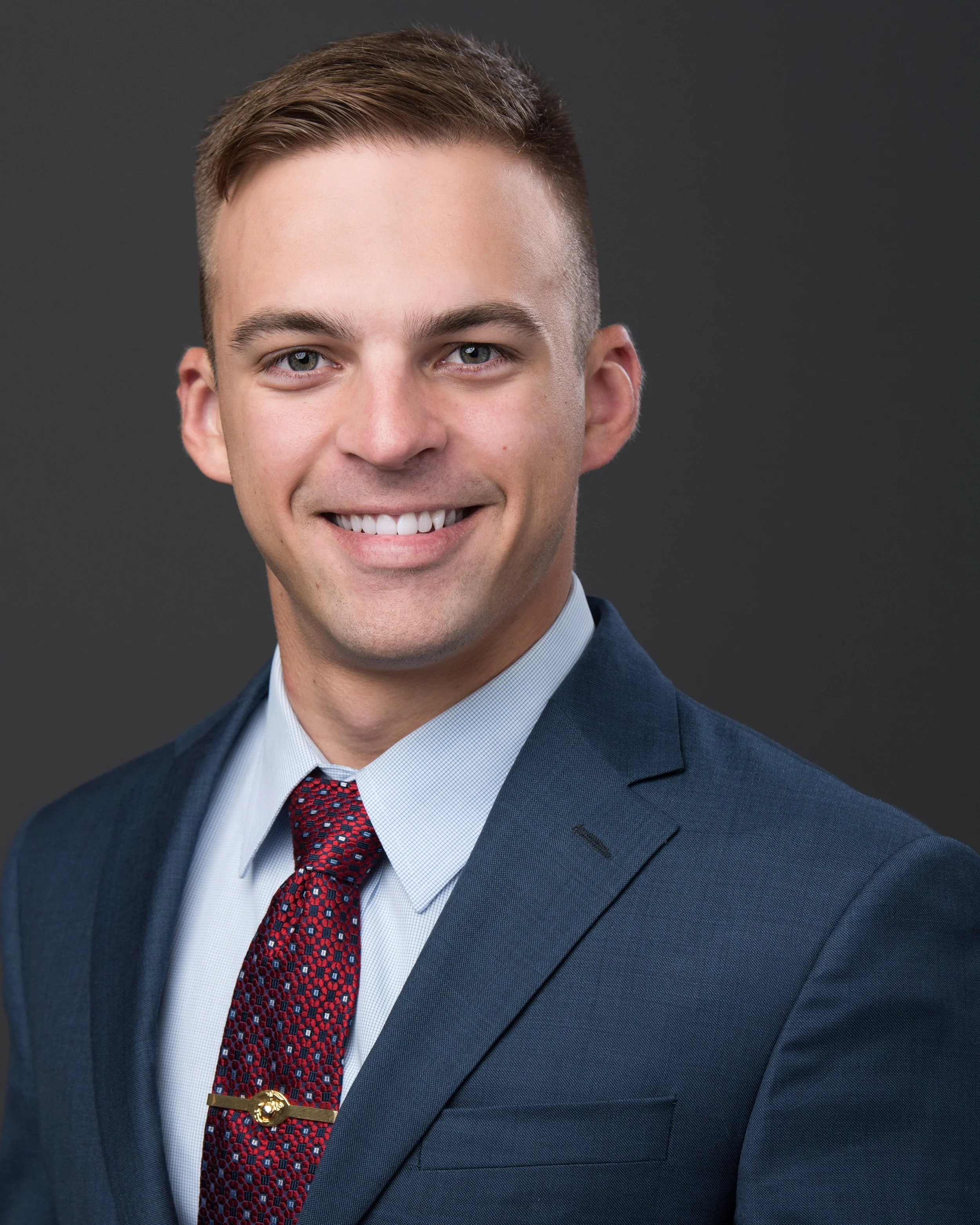 Jared Plamann headhot. He is wearing a dark blue jacket, light blue shirt, red patterned tie, and a gold tie clip.