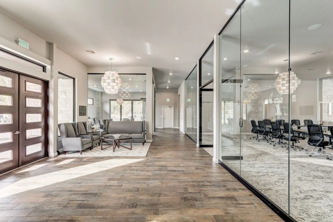 Modern interior space with a seating area and a conference room separated by glass walls, wooden flooring, and contemporary lighting fixtures.