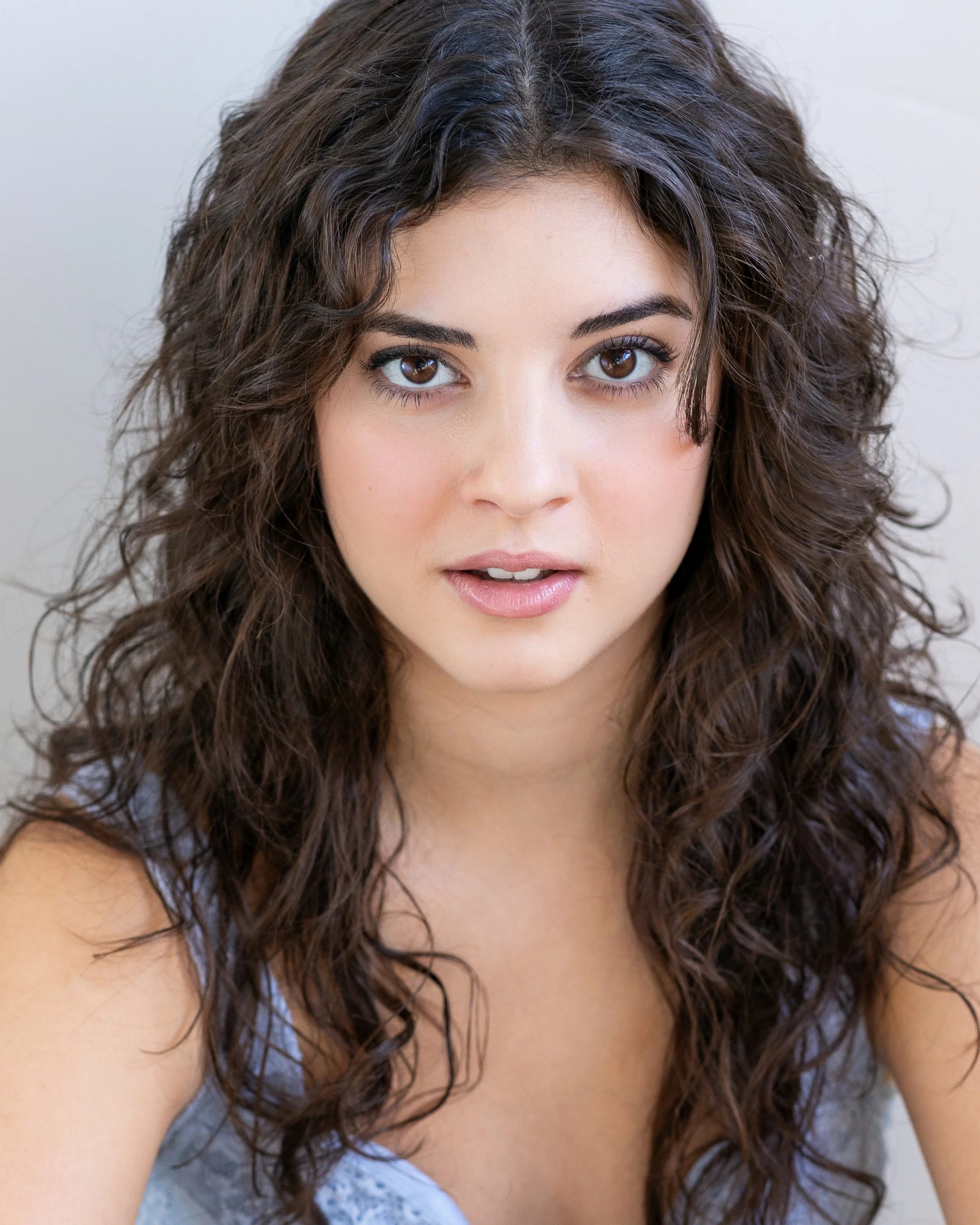 Close-up portrait of a young woman with wavy dark brown hair, brown eyes, and a subtle smile.