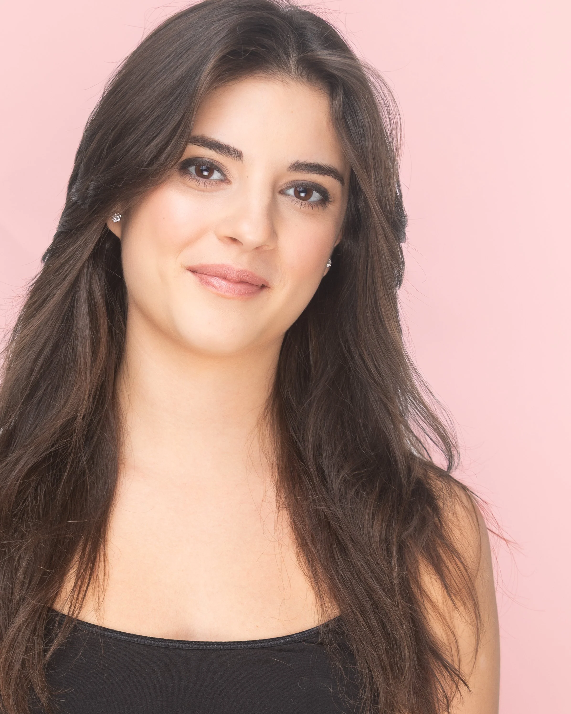 A young woman with long brown hair wearing a black top, smiling softly, against a light pink background.