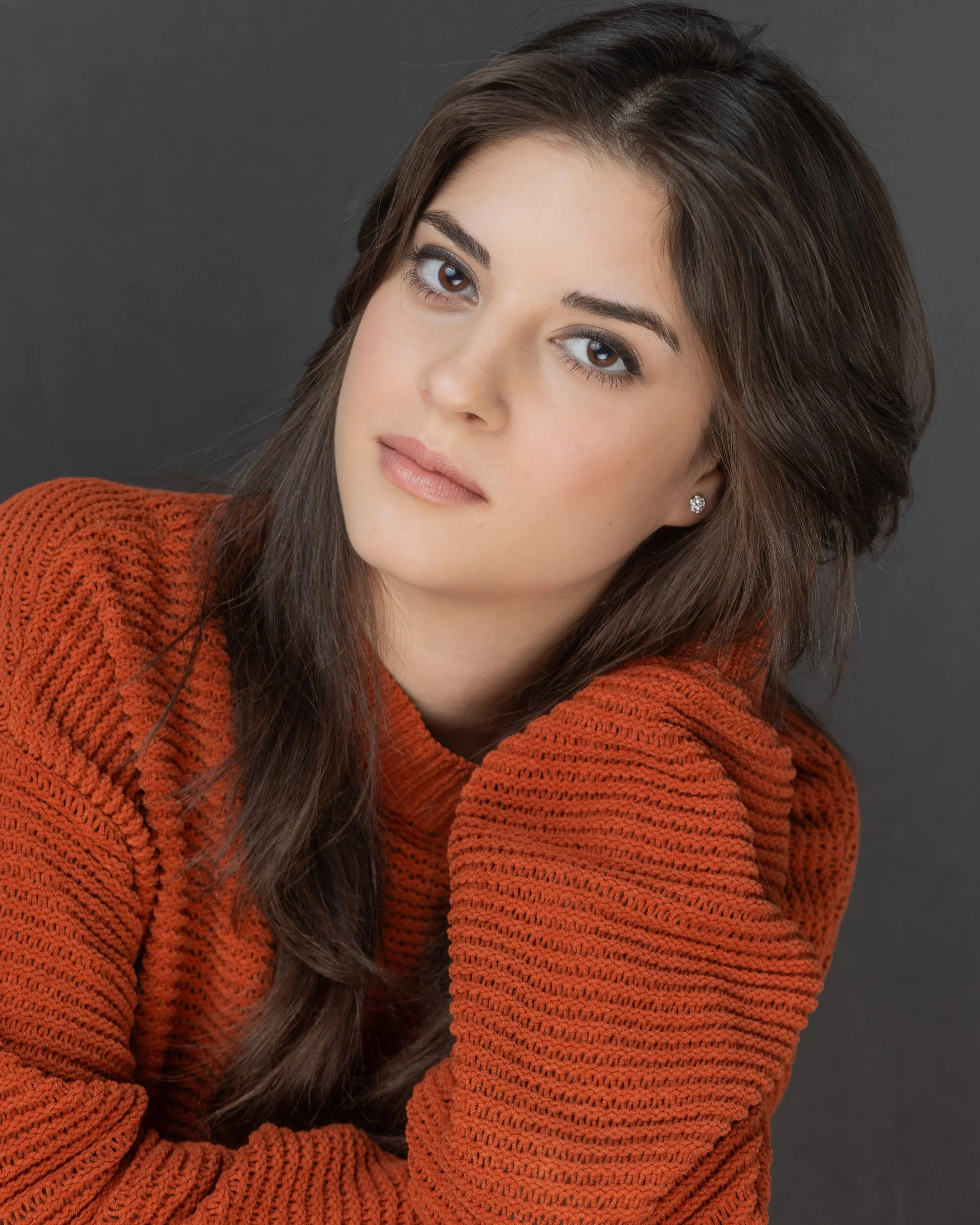 Close-up portrait of a young woman with brown hair, blue eyes, wearing a rust-colored knitted sweater, against a dark gray background.