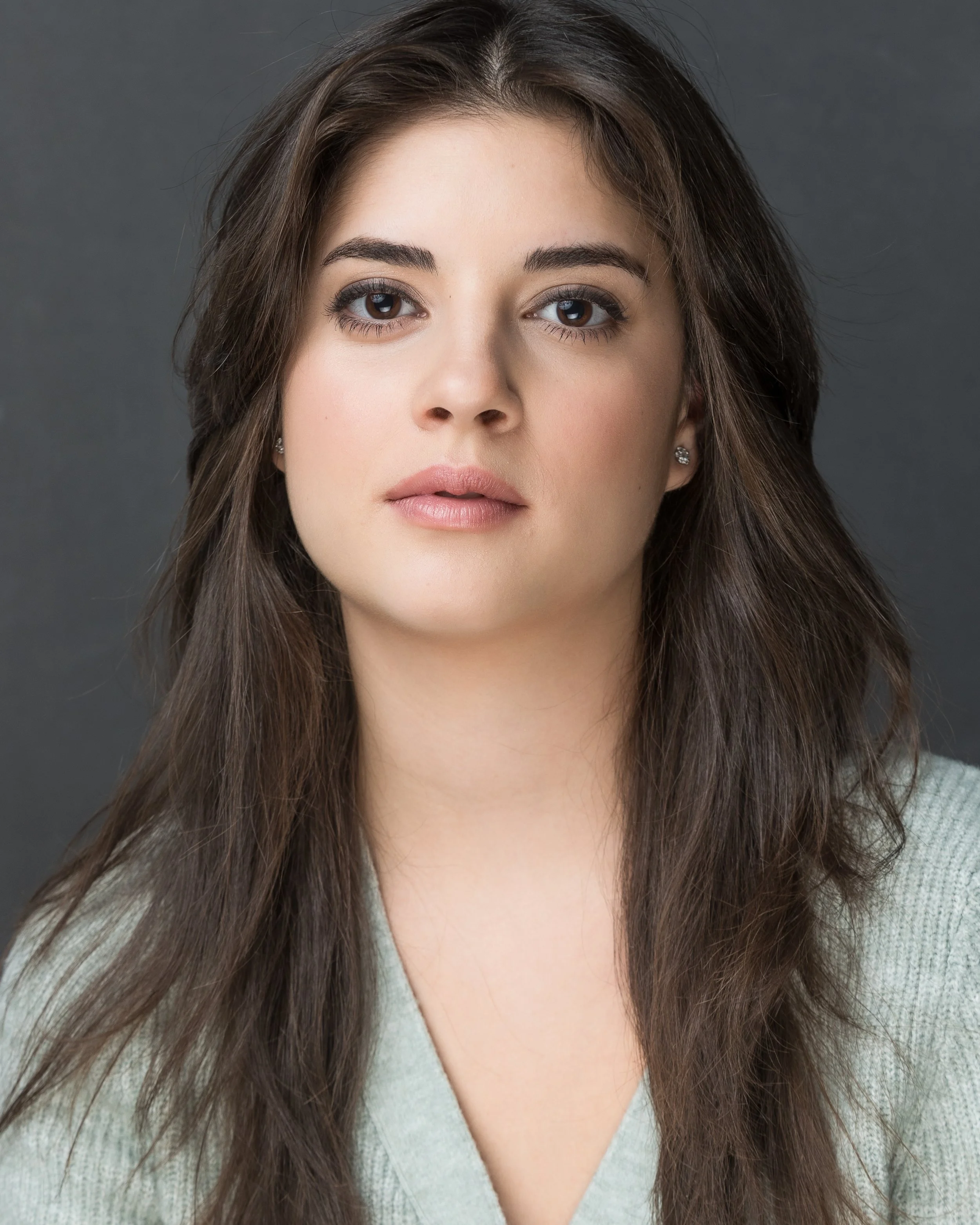 Close-up portrait of a young woman with brown hair and fair skin, wearing a light-colored top, against a dark gray background.