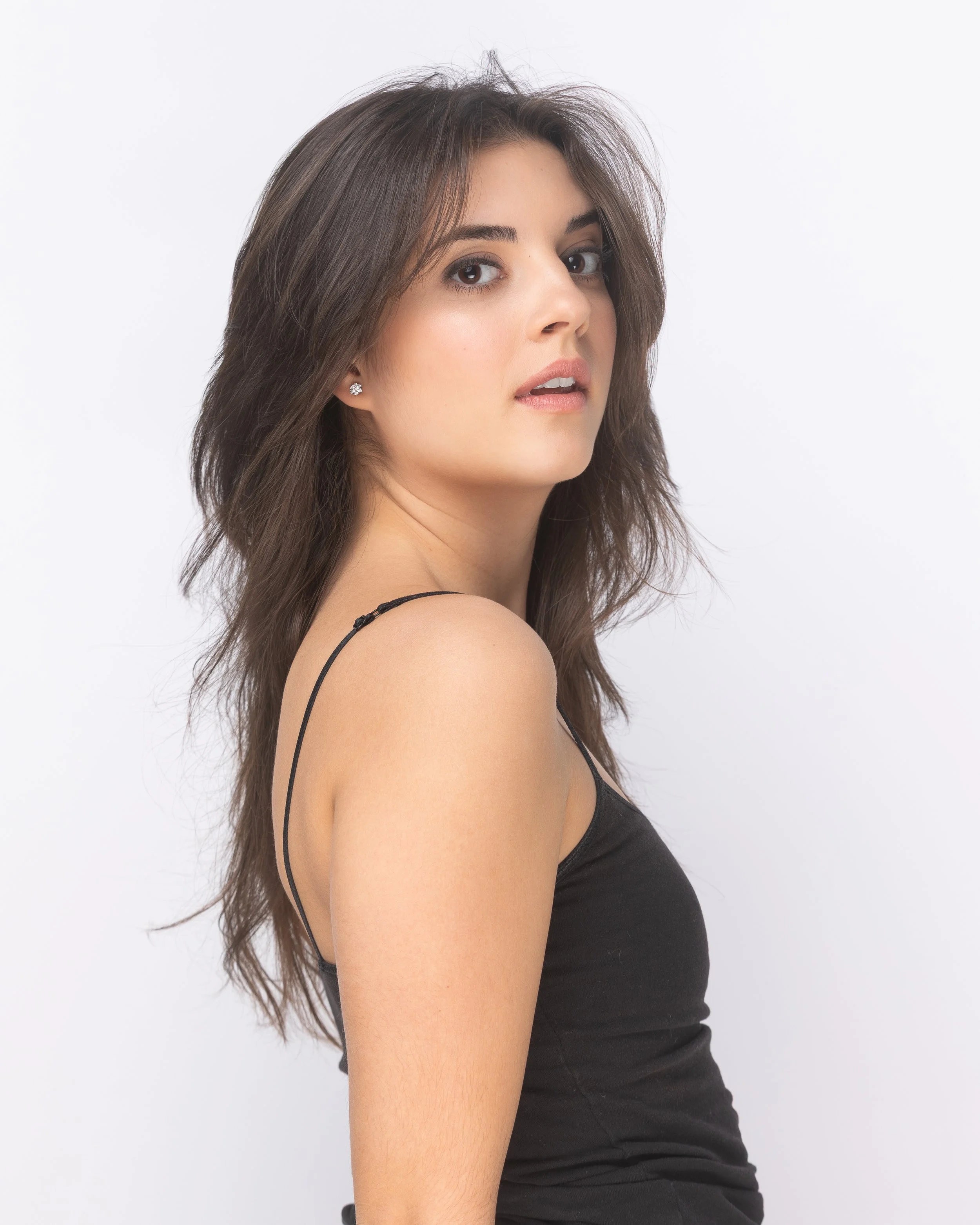 A woman with long, wavy brown hair posing with her head turned slightly to the side against a plain white background.