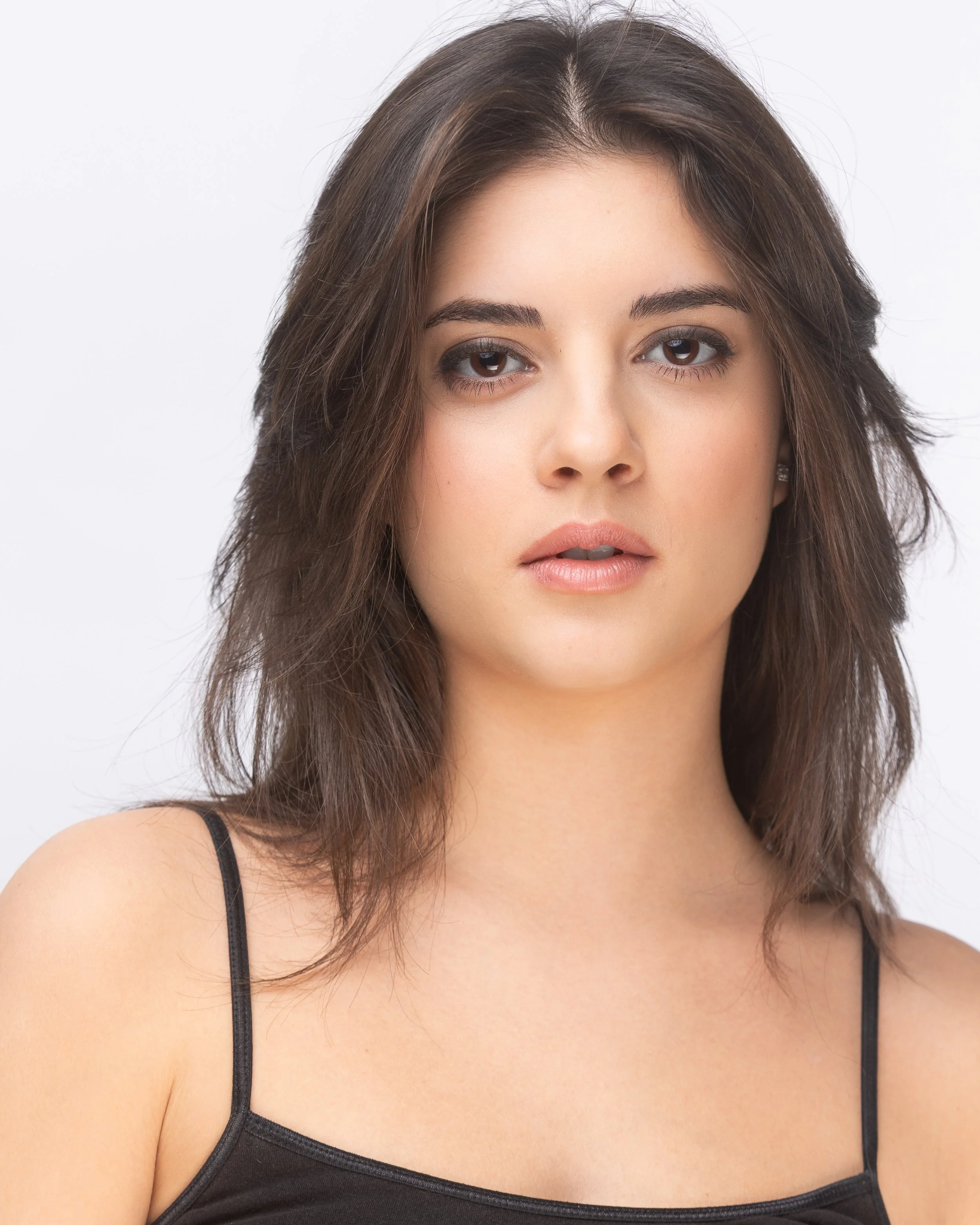 A young woman with shoulder-length brown hair, neutral makeup, and wearing a black spaghetti strap top, looking directly at the camera against a plain light background.
