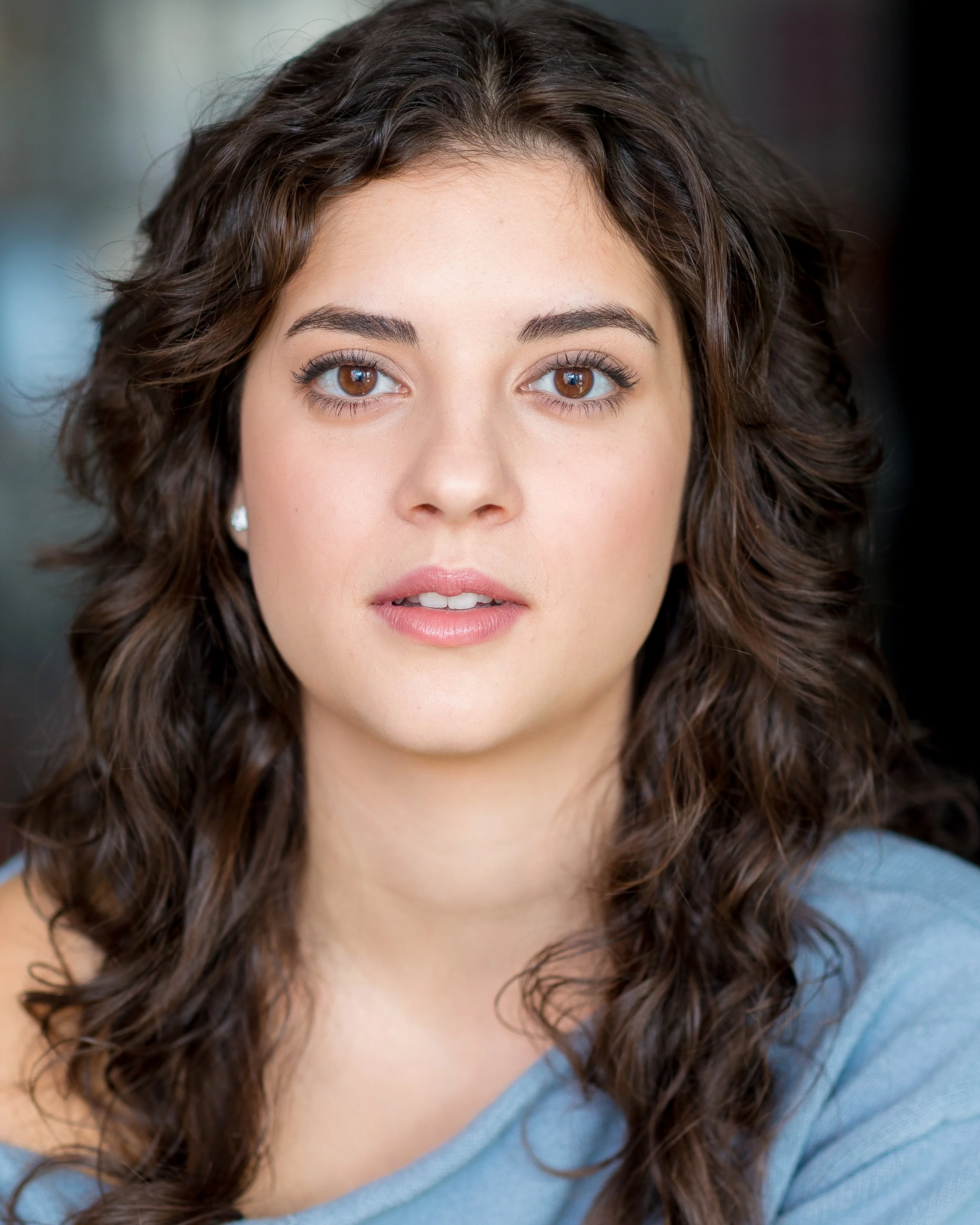 Close-up of a young woman with curly brown hair and brown eyes, wearing a light blue top and small earrings, looking directly at the camera.