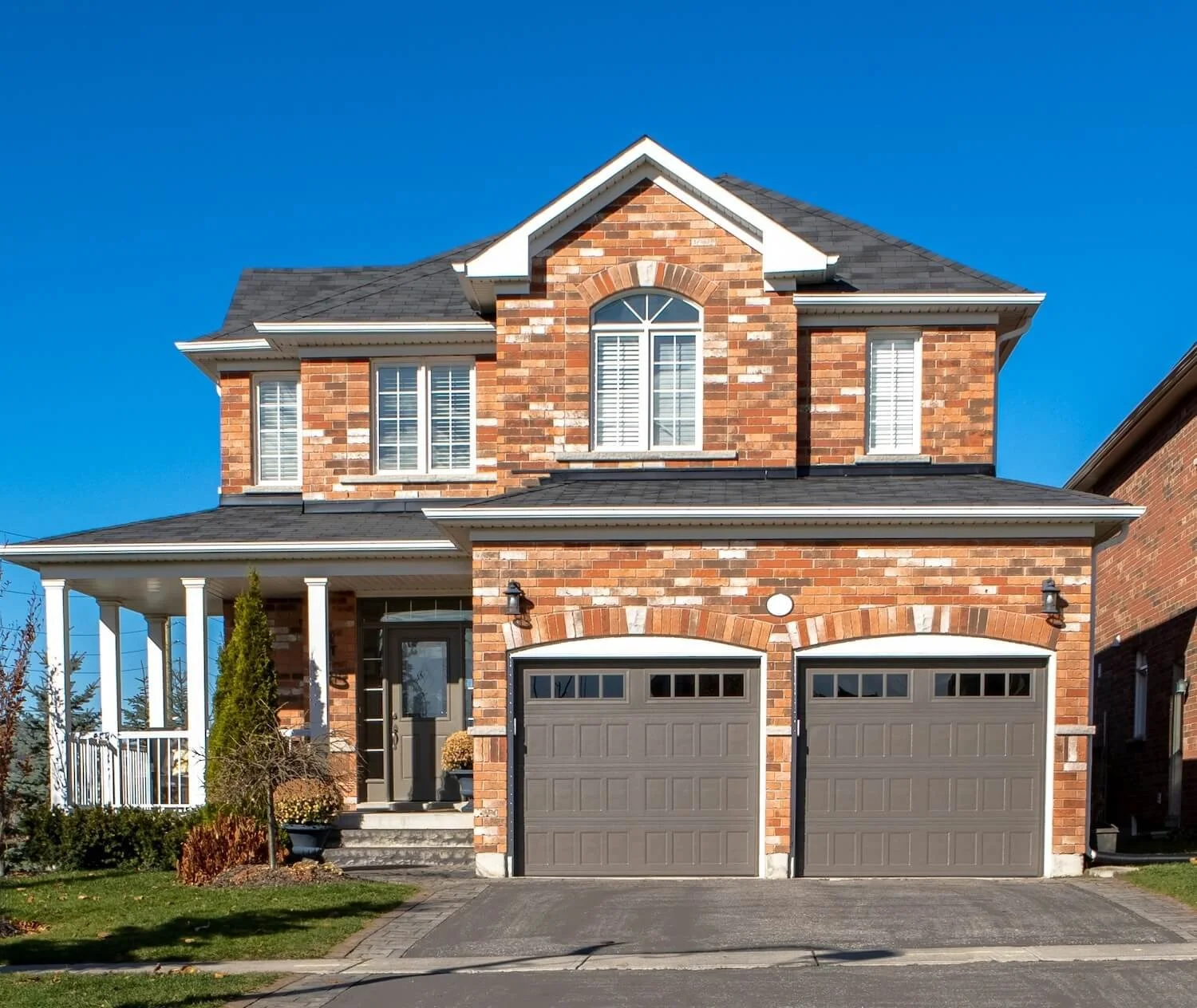 A two-story brick house with a black shingle roof, white window trims, two garage doors, and a front porch with white columns under a clear blue sky.
