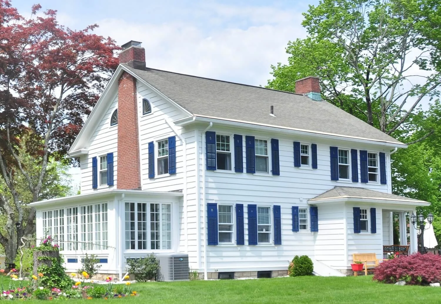 A two-story white house with blue shutters, brick chimneys, and a porch, surrounded by green trees and a well-maintained lawn with flowers.