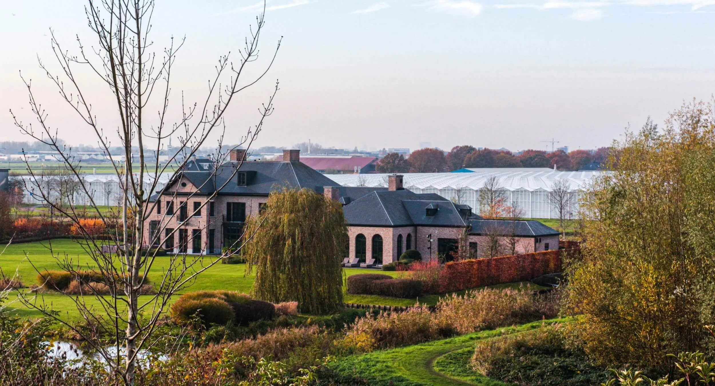 A large house with a green lawn and trees, with greenhouses in the background
