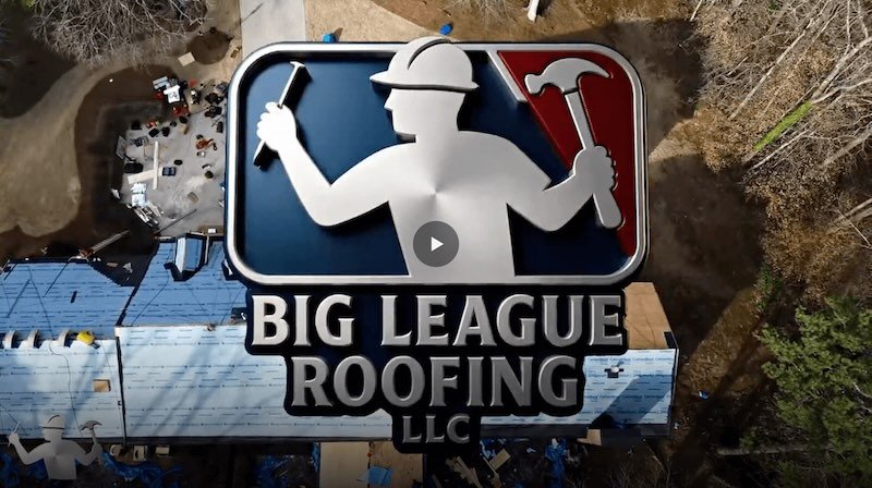 A sign for Big League Roofing LLC with a silhouetted roof worker holding a hammer and a nail, featuring the logo's red and blue background, mounted on a building under construction.