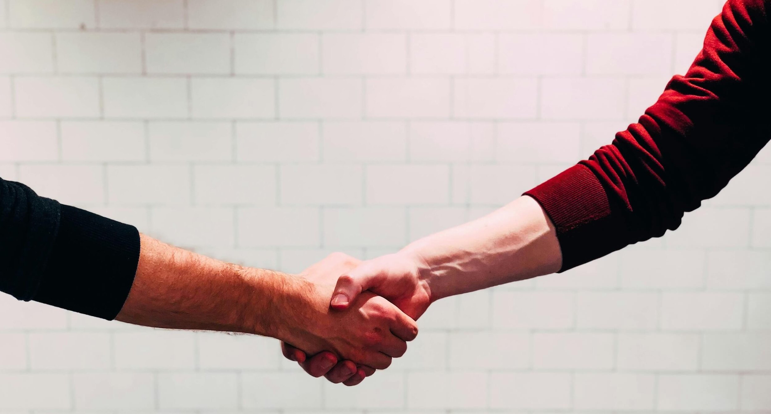 Two individuals shaking hands in front of a white brick wall.