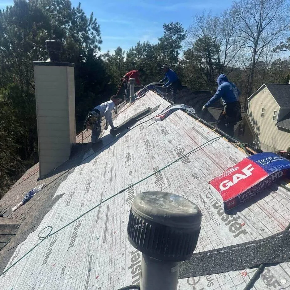 Workers installing or repairing a roof on a residential house during daytime, with roofing materials and tools visible.