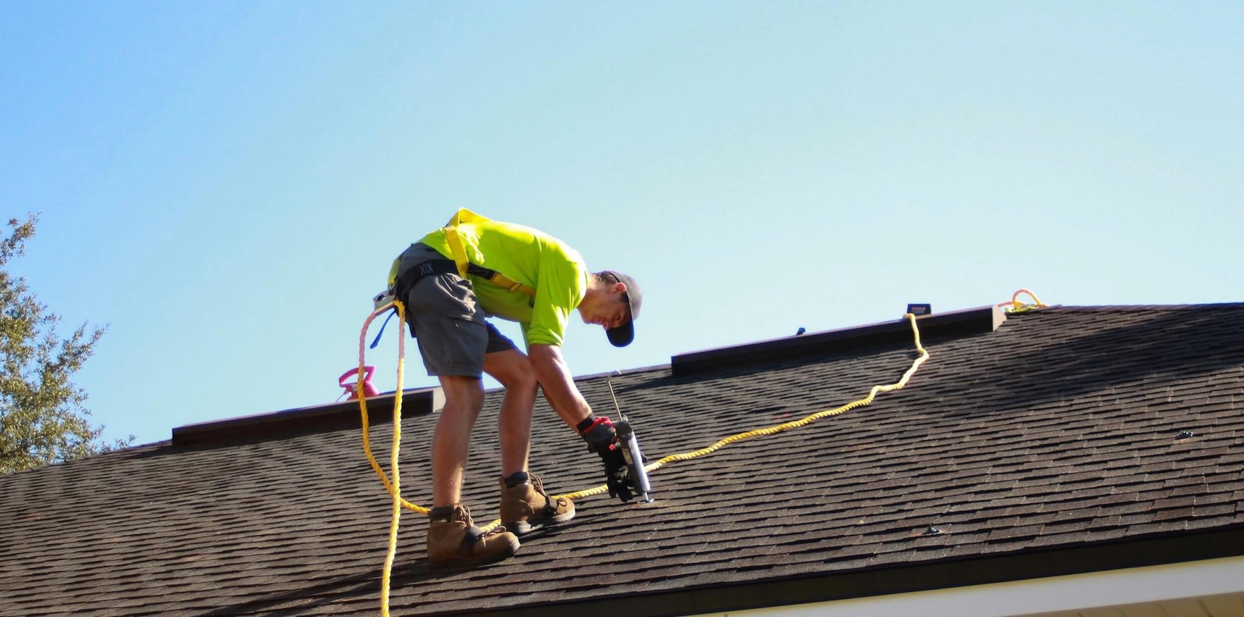 A man wearing a bright yellow safety jacket, gray shorts, and brown work boots is repairing a roof while standing on its sloped surface, secured with a safety harness and a yellow rope.
