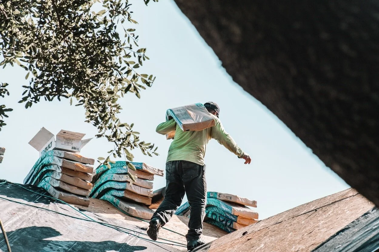 A person carrying a bag of supplies walking on a rooftop with stacks of bags nearby, under a clear sky with tree branches overhead.