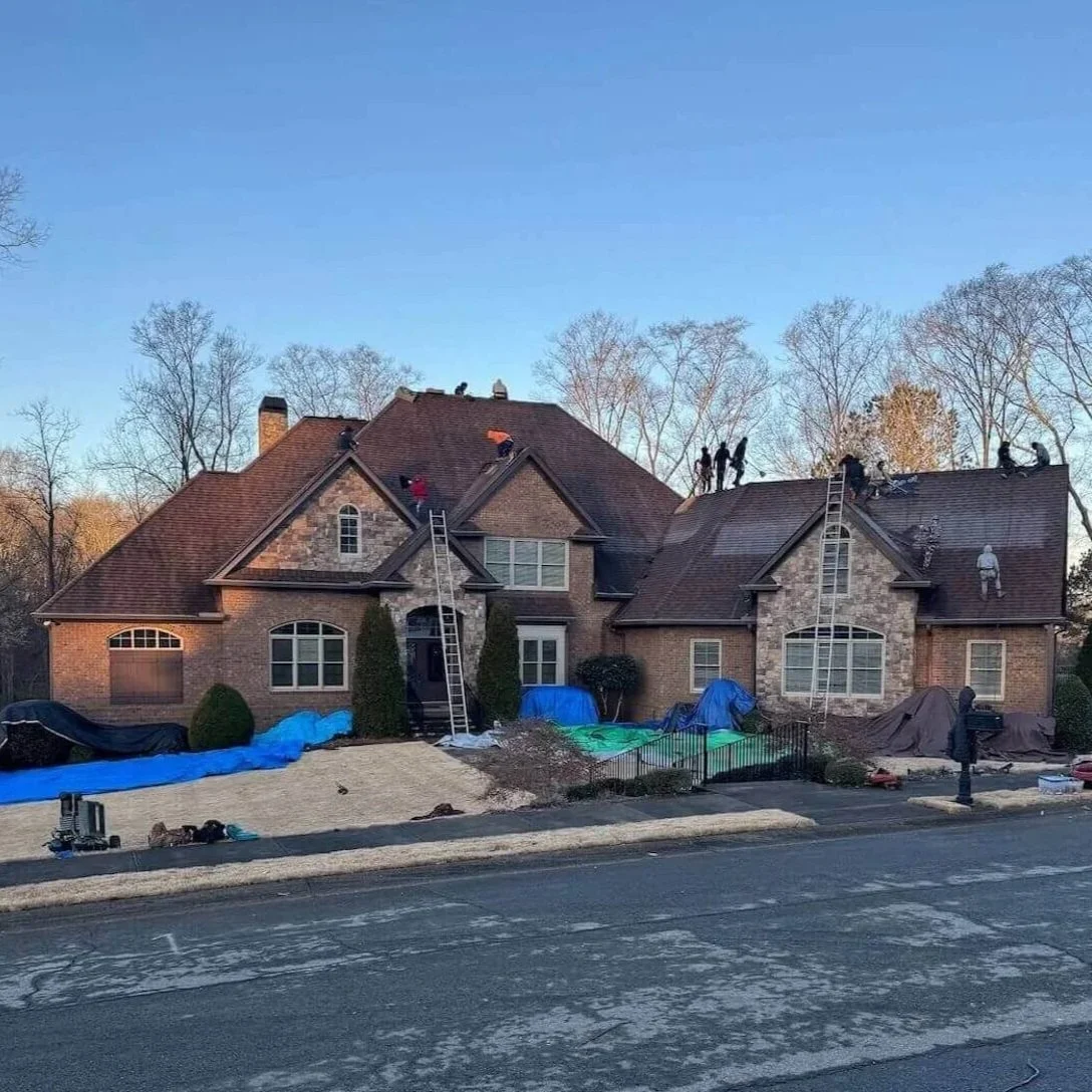 Construction workers on the roof of a brick house with ladders and tools, working on roof repairs or replacement on a clear day.