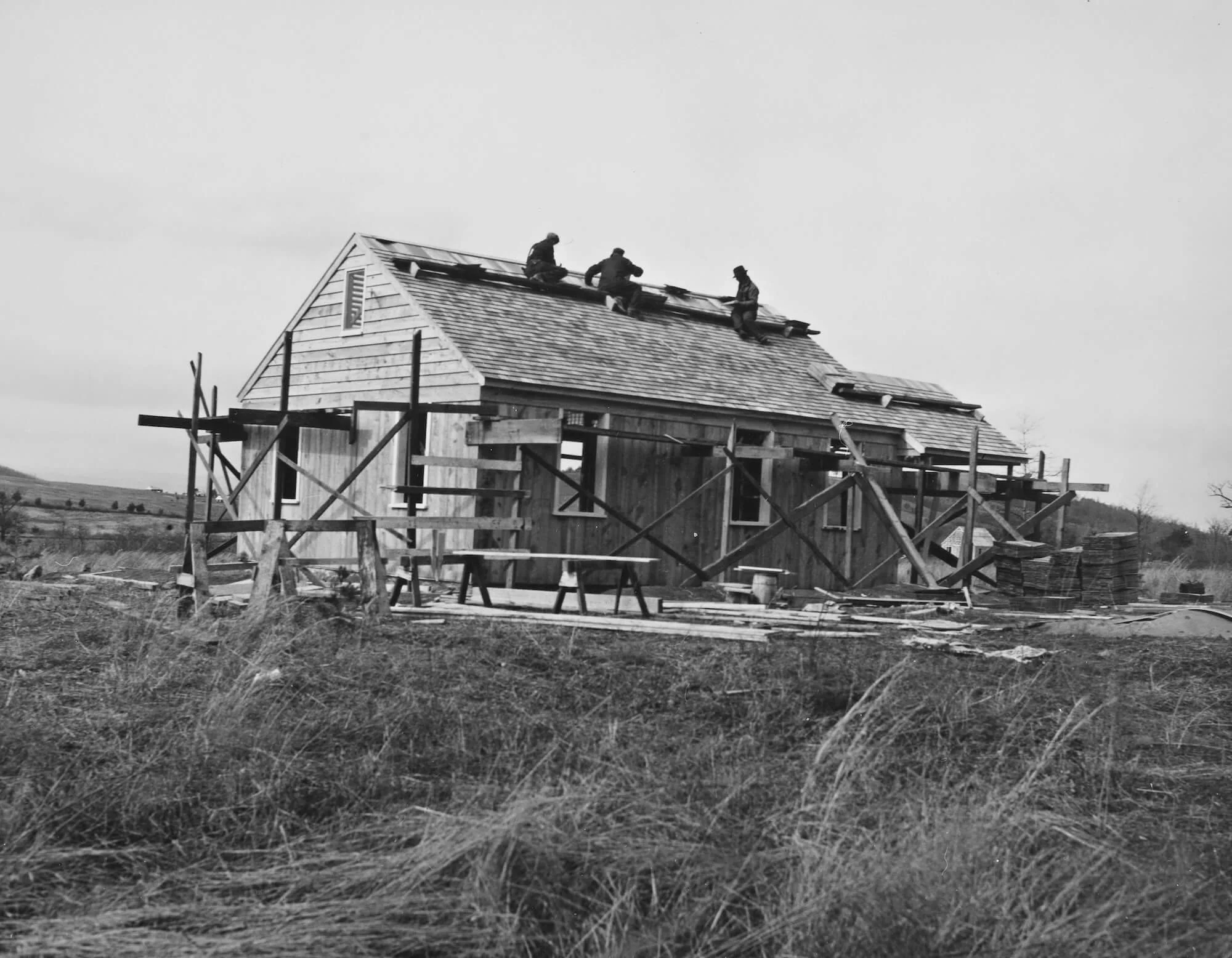 Black and white photo of a house under construction with scaffolding around it. Three workers are on the roof working on the shingles.