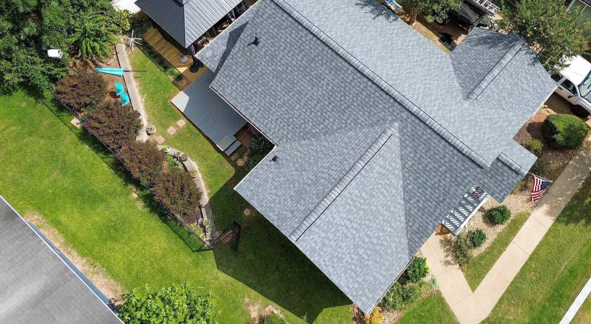 Aerial view of a house with gray shingle roof, front porch, and landscaped yard with green lawn, bushes, trees, a swing, and a small patio area. A sidewalk runs along the front and side of the property, and there is a parked white vehicle nearby.