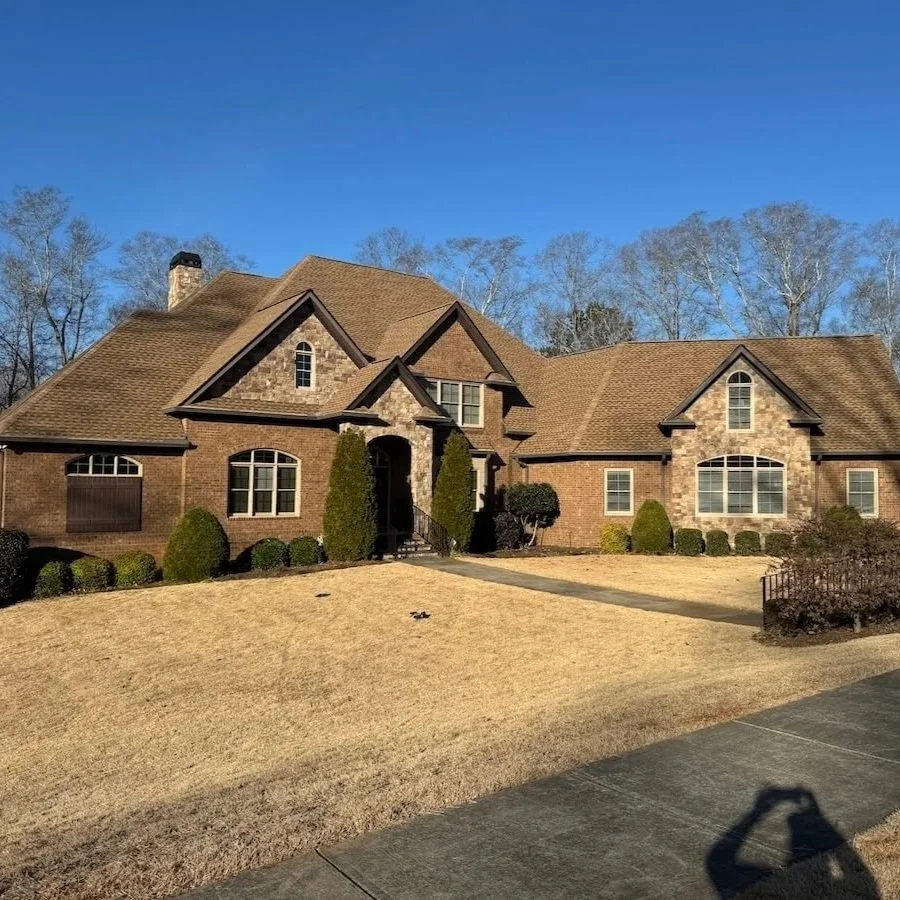 A large two-story brick house with a brown shingle roof, multiple gables, and a chimney. The front yard has dry, yellow grass, a concrete walkway leading to the front door, and landscaped bushes and trees.