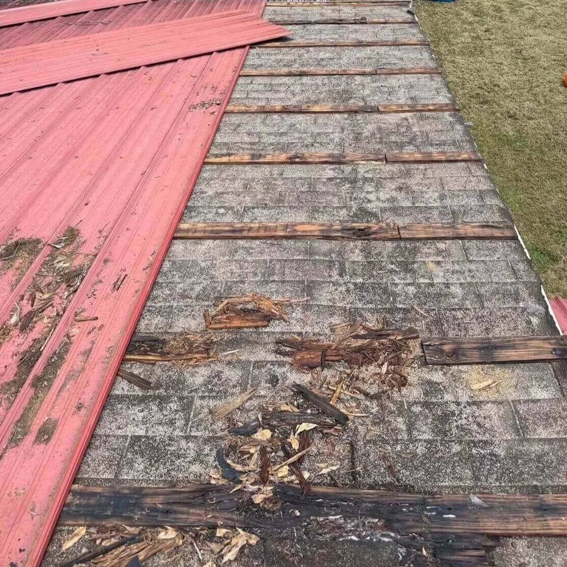 Part of a roof with damaged shingles and siding missing, showing the underlying wood structure and debris, with dirt and leaves. The shingles are aged and weathered.