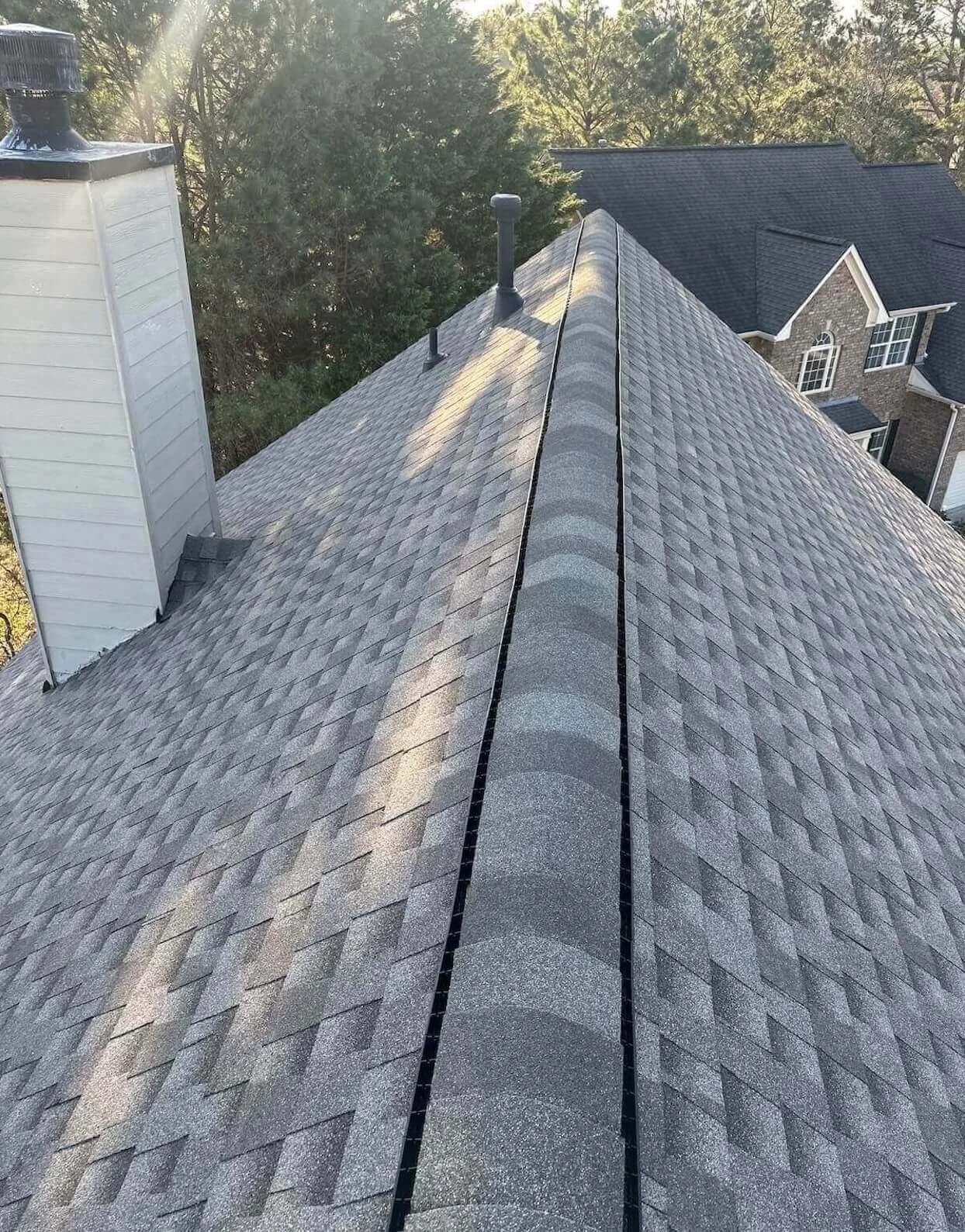A view of a house's gray shingle roof, showing vents and chimney, with trees and a neighboring house in the background.