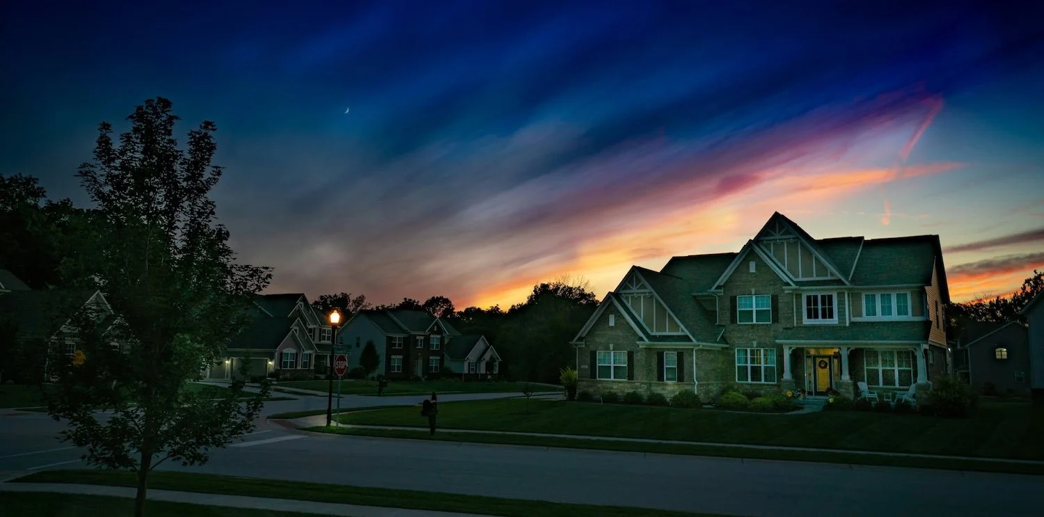Residential neighborhood during sunset with houses, trees, streetlamp, stop sign, and a person walking on the sidewalk.