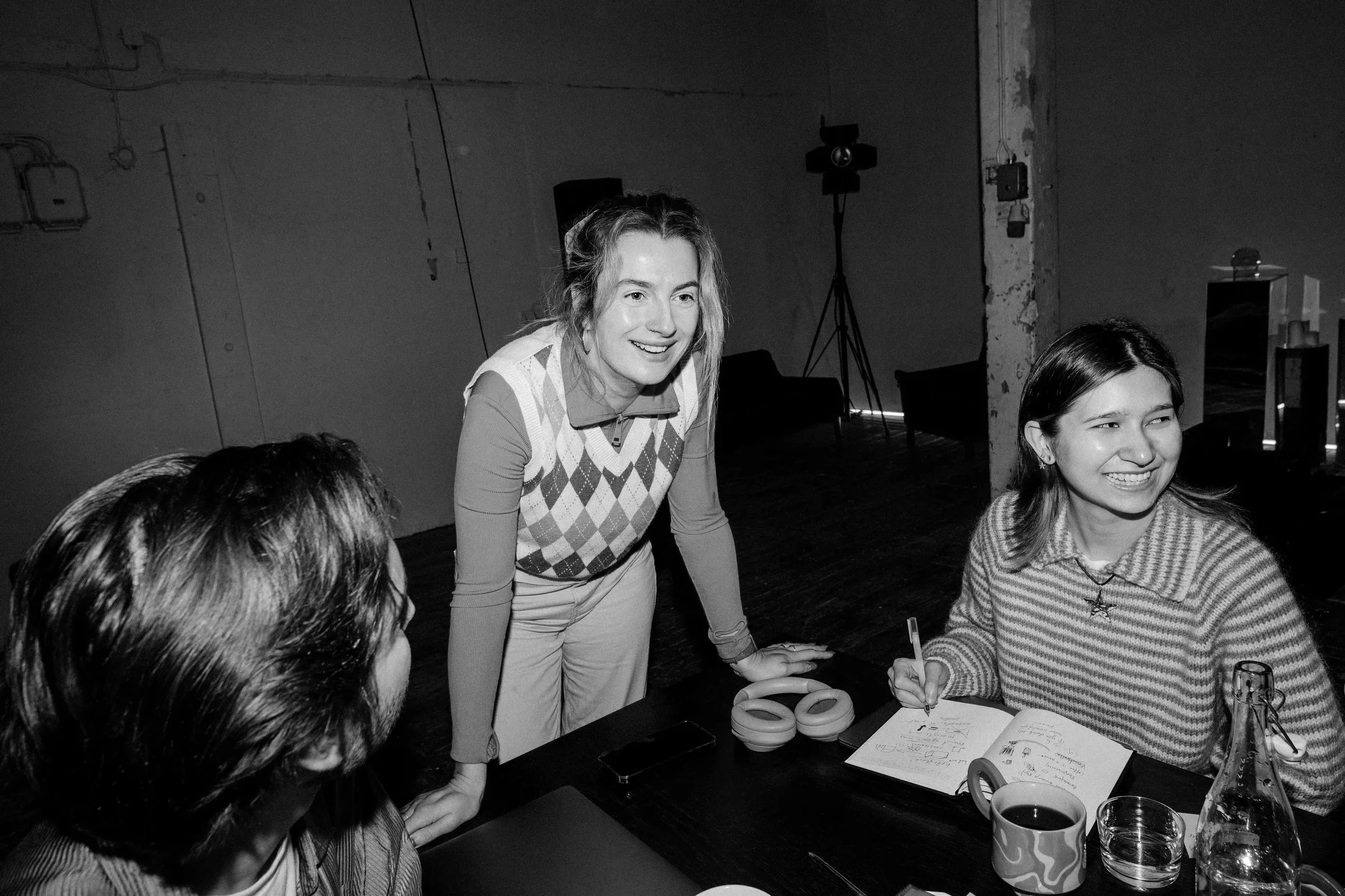 Une femme souriante en veston karaté blanc avec un motif argyle sur le devant, debout à une table, entourée de plusieurs jeunes femmes assises qui prennent des notes ou discutent, dans une pièce avec un fond industriel, éclairée par des lumières, ambiance of style workshop ou réunion informelle.