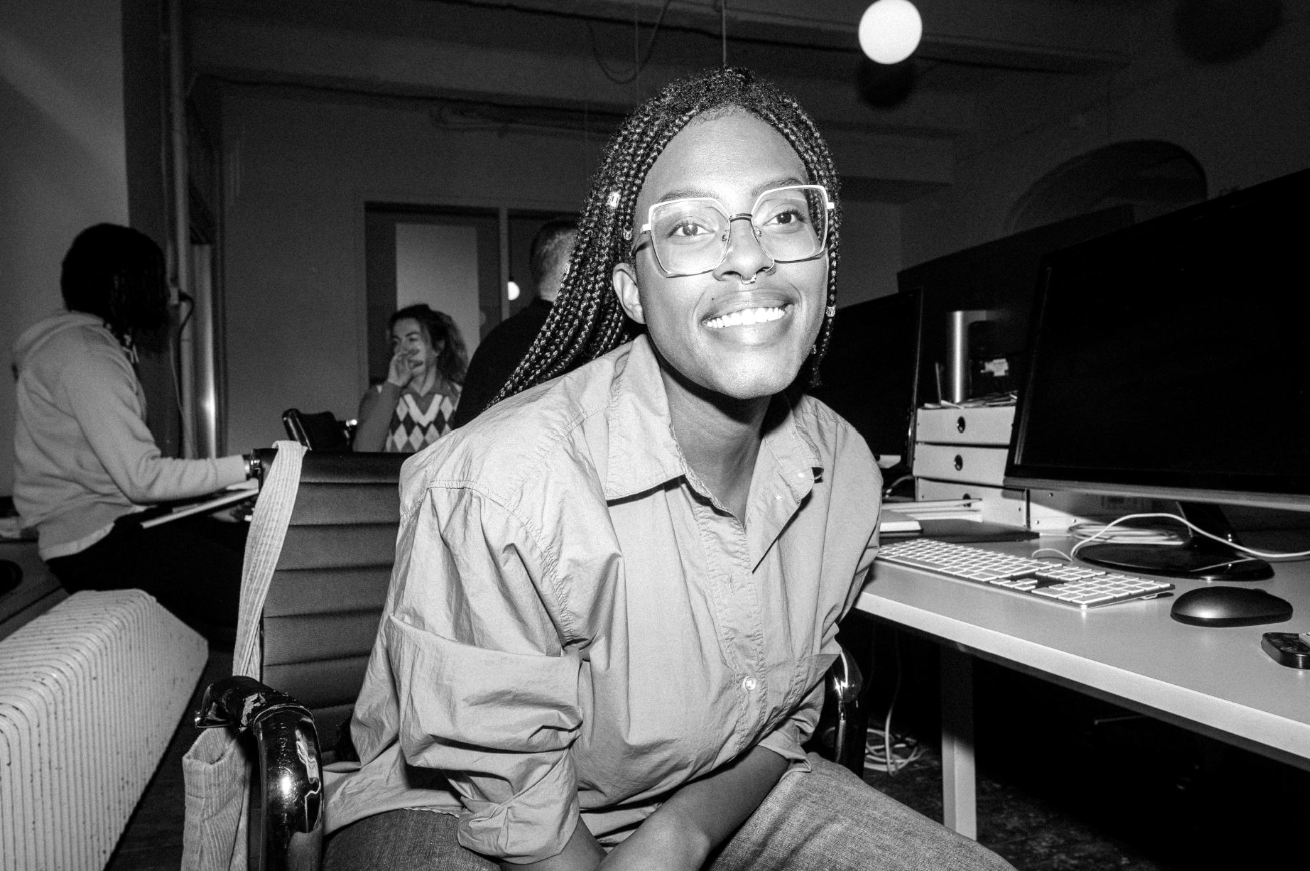 Jeune femme souriante assise devant un ordinateur dans un environnement de bureau ou de coworking, autres personnes en arrière-plan.