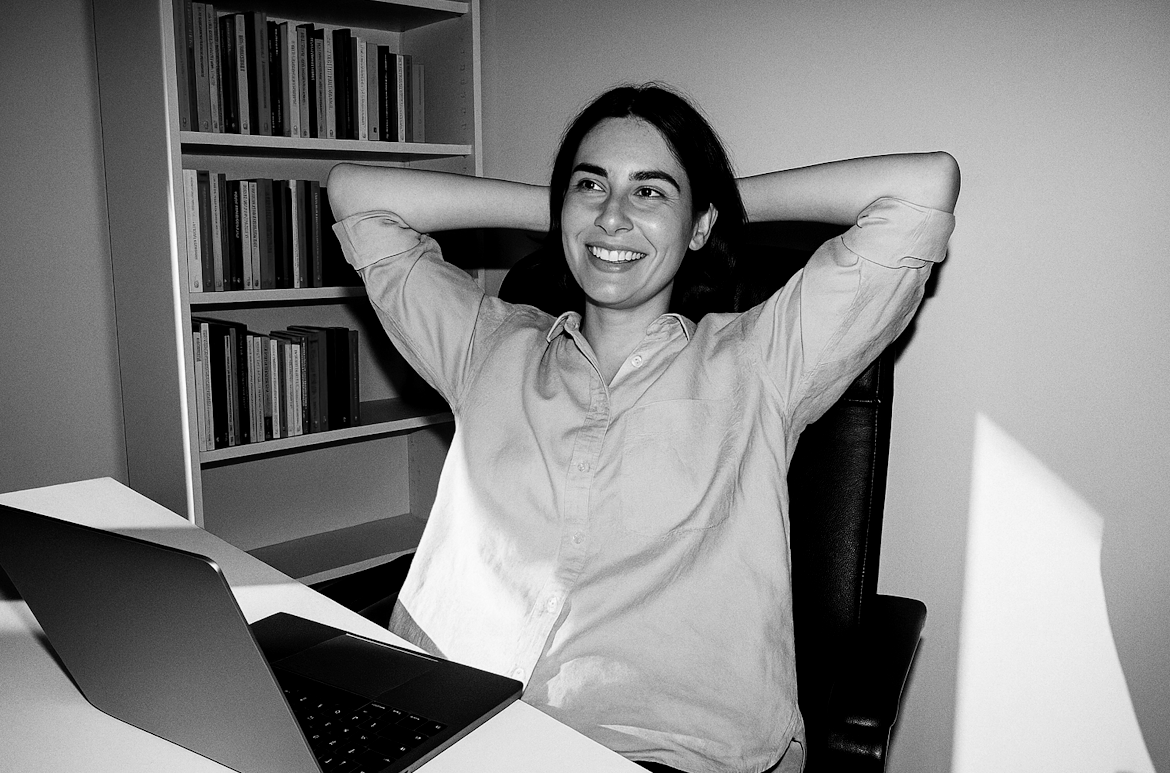 Une femme souriante, assise à son bureau avec un ordinateur portable, dans un environnement de bureau, en noir et blanc.