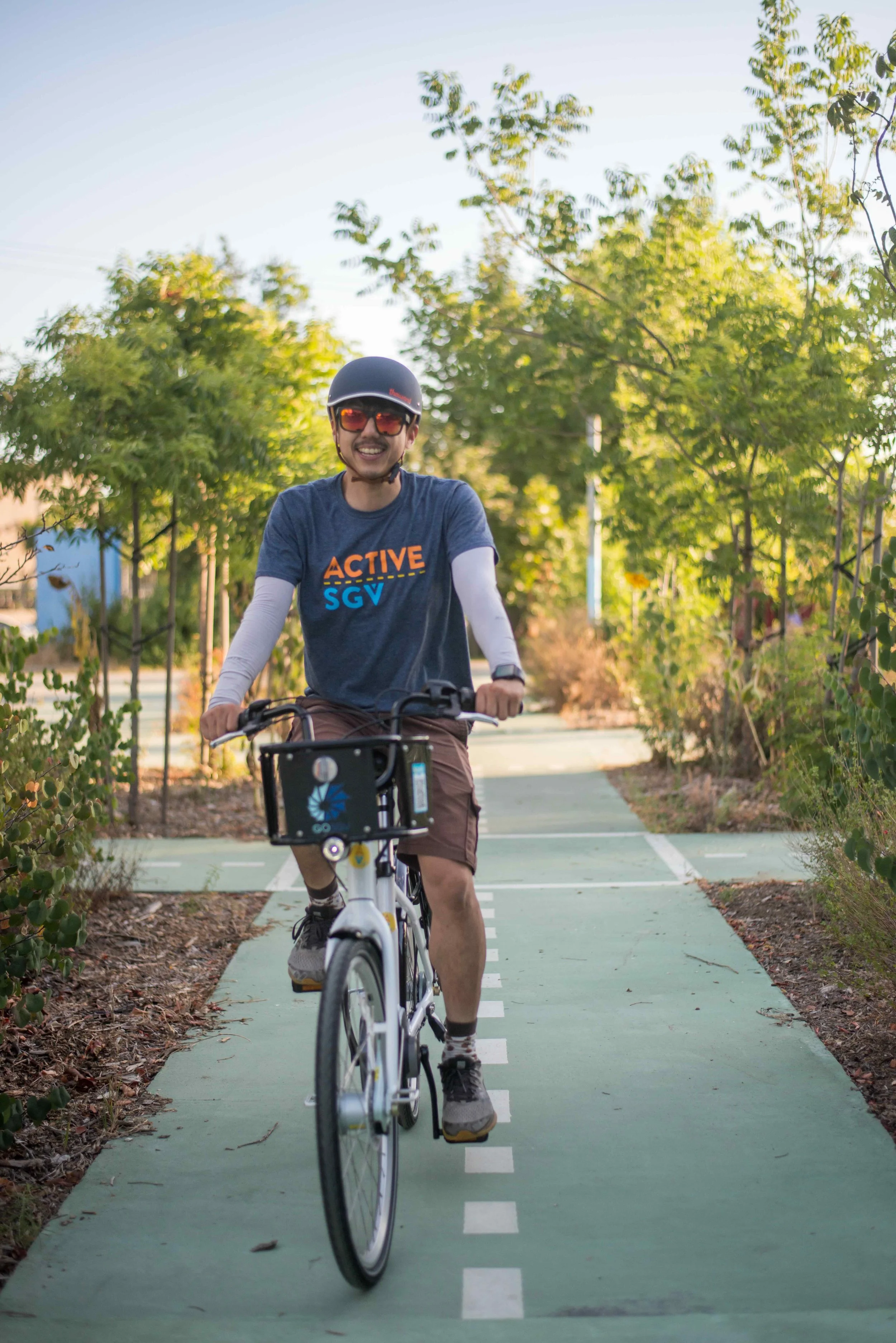 Man riding a bicycle on a designated bike lane surrounded by trees, wearing a helmet, sunglasses, and a dark T-shirt with 'ACTIVE SGV' logo.