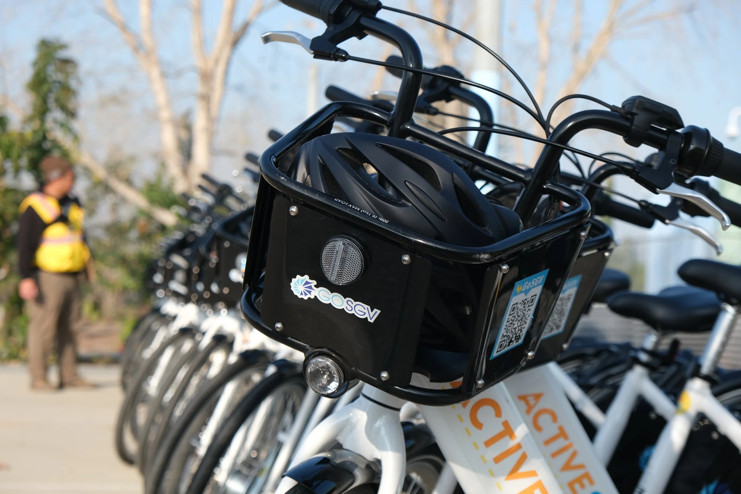 Row of white bikes with black baskets containing helmets parked outdoors, with a person wearing a yellow vest and glasses standing in the background.