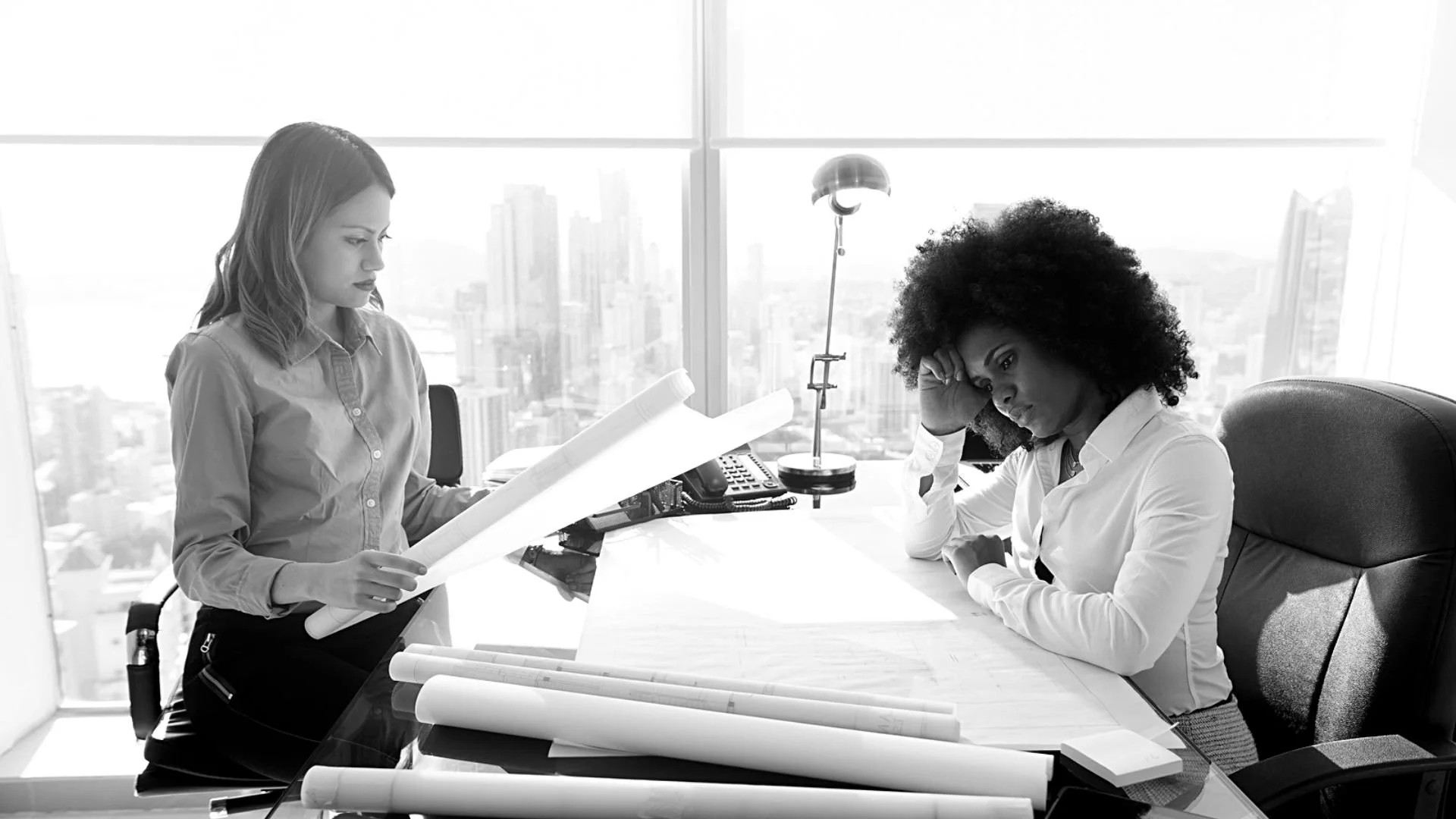Two women in an office with large windows, one is standing and reading documents, the other is sitting with her hand on her forehead, looking at the desk with rolled-up papers.