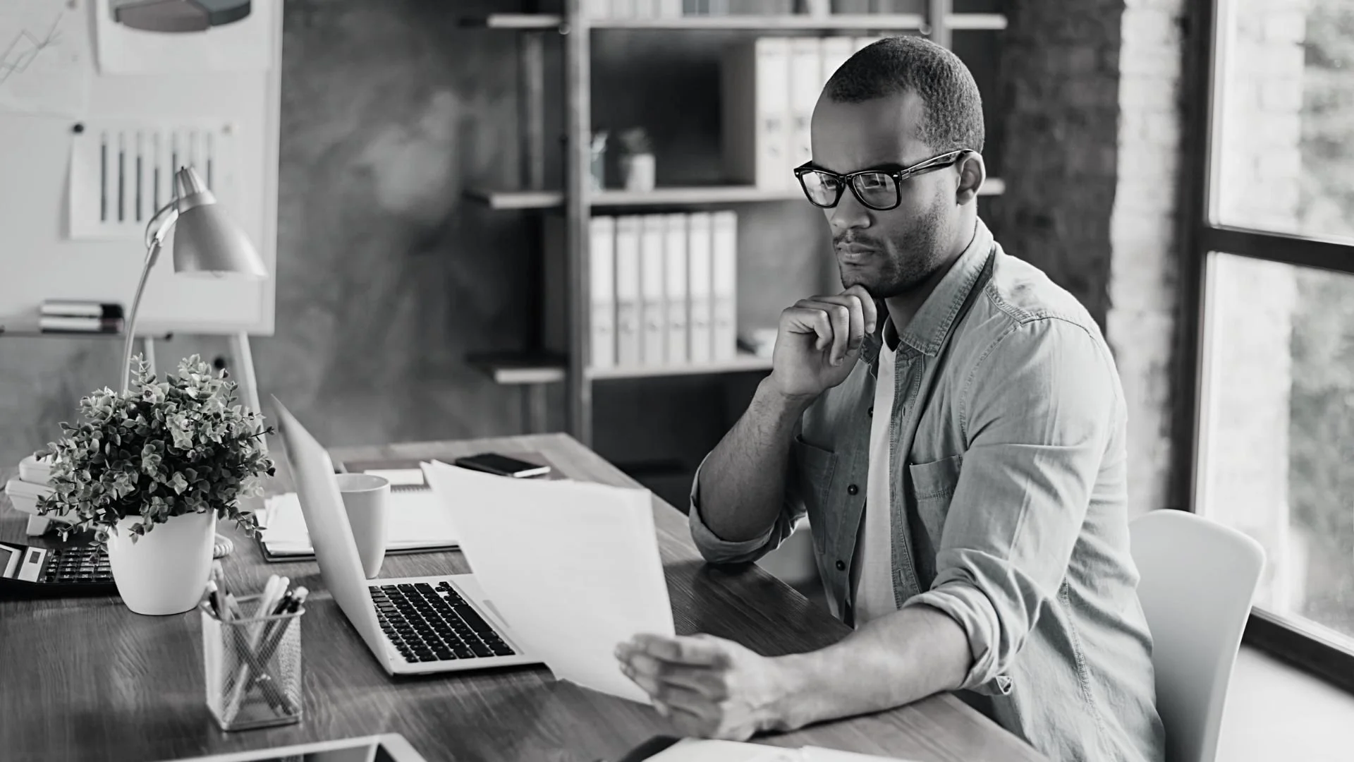 A man wearing glasses working at a desk with an open laptop, papers, and office supplies, in a well-lit office with large windows.