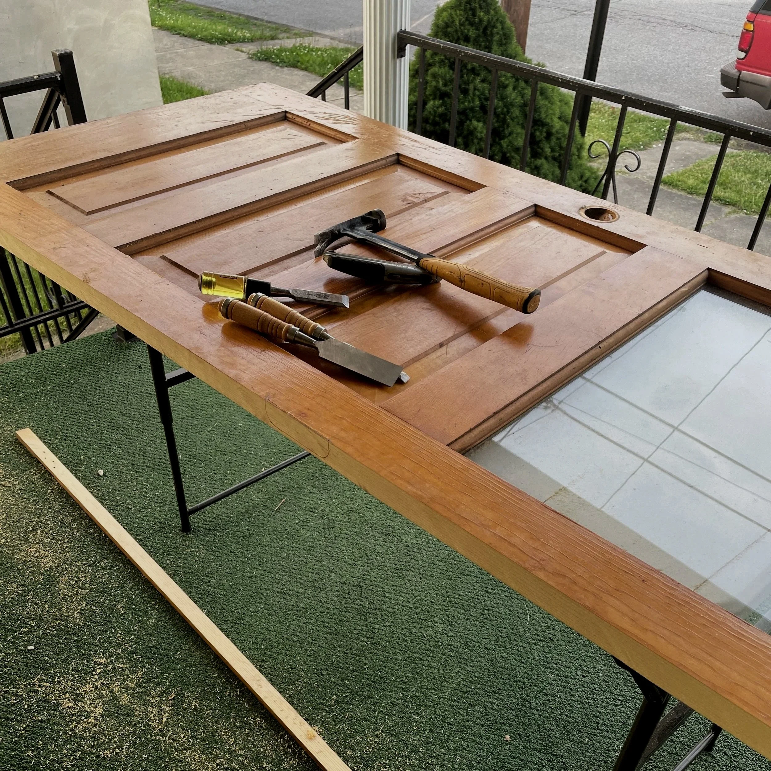 Wooden door panel on a table with tools including a hammer, chisels, and a wood strip, outdoors on a porch.