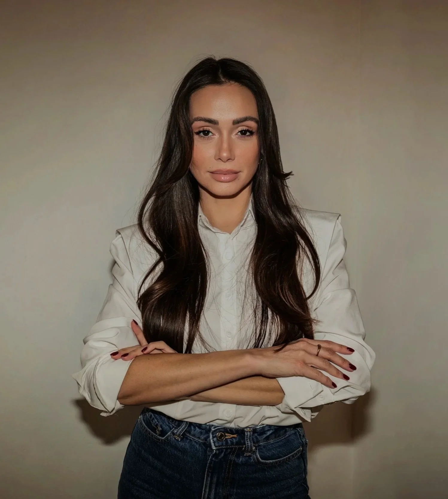 Junge Frau mit langen braunen Haaren, weißem Hemd und Jeans vor neutralem Hintergrund.