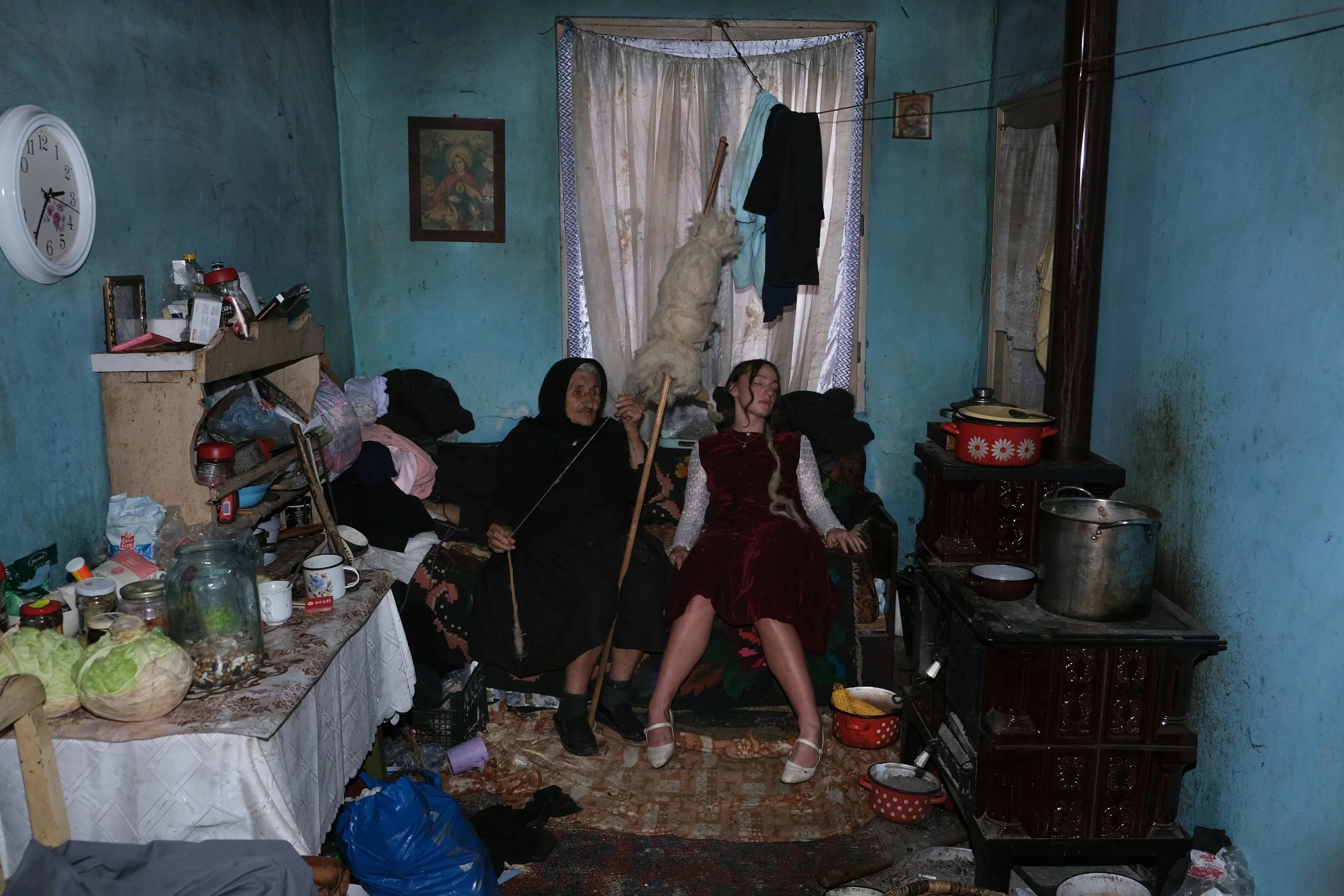 Inside a cluttered living room with blue walls, two women sit on a sofa. An elderly woman dressed in black holds a stick, and a younger woman in a red dress and heels sits with eyes closed. Items such as jars, food, and clothes are scattered around the room, with a stove and pots on the right and a table with food and containers on the left.