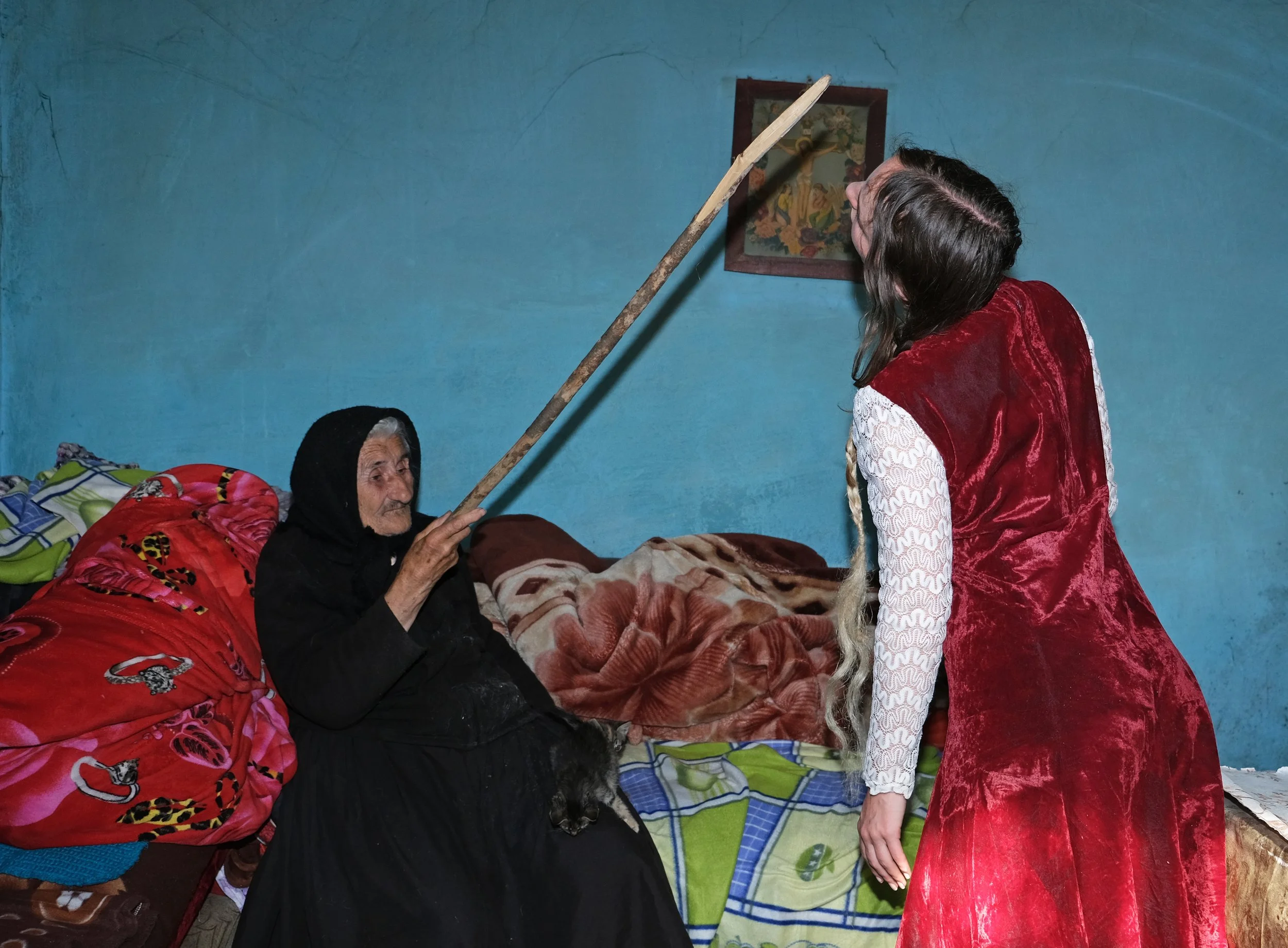 An elderly woman sitting in bed, holding a stick, with a young woman dressed in a red dress, inside a modest bedroom with a blue wall.
