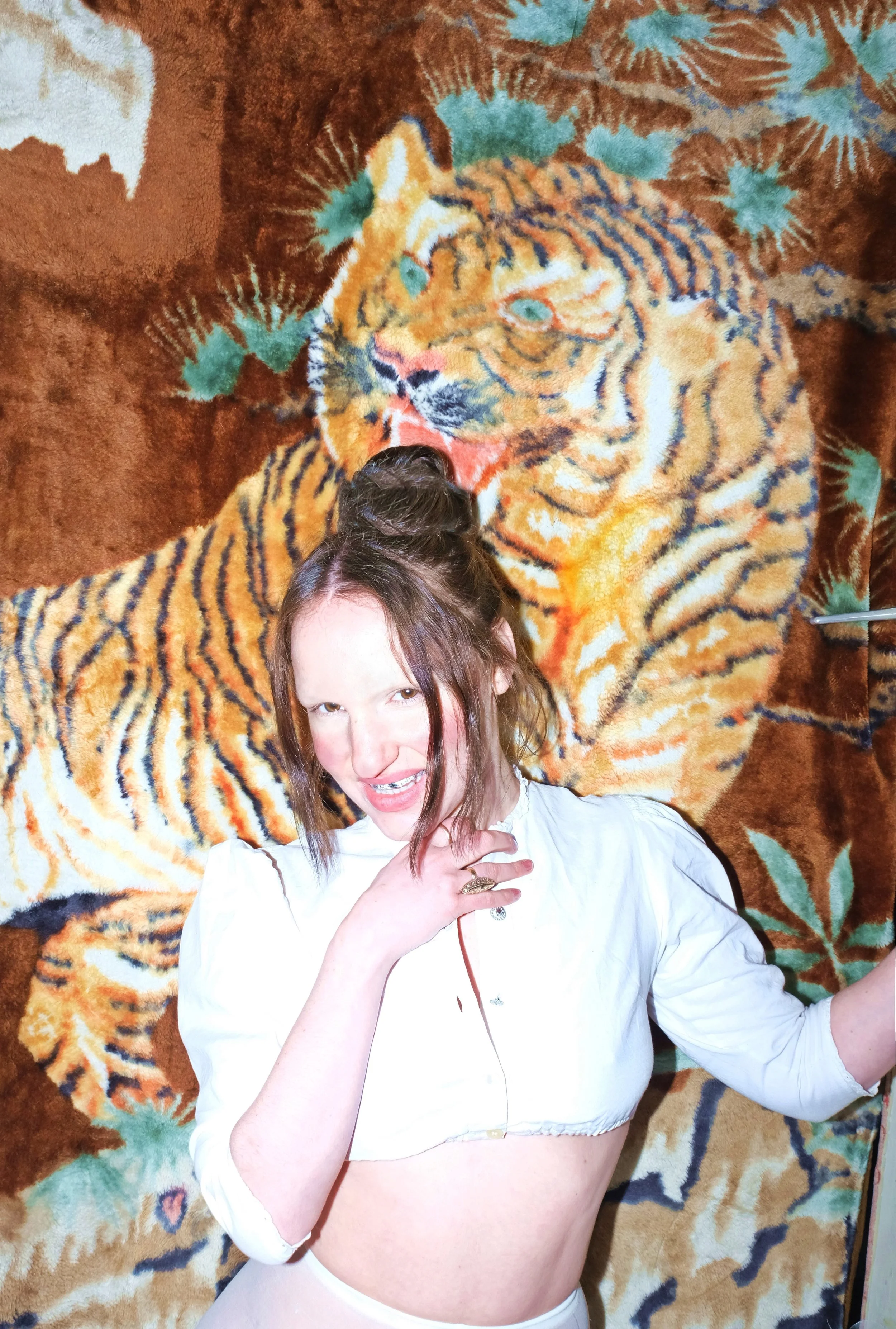 A woman with light skin, brown hair styled in a top knot, posing in front of a large tiger-themed textile or tapestry. She is wearing a white, cropped, long-sleeve top with puffed shoulders and a ruffled neckline, and has a playful expression with her mouth open and her hand near her neck.