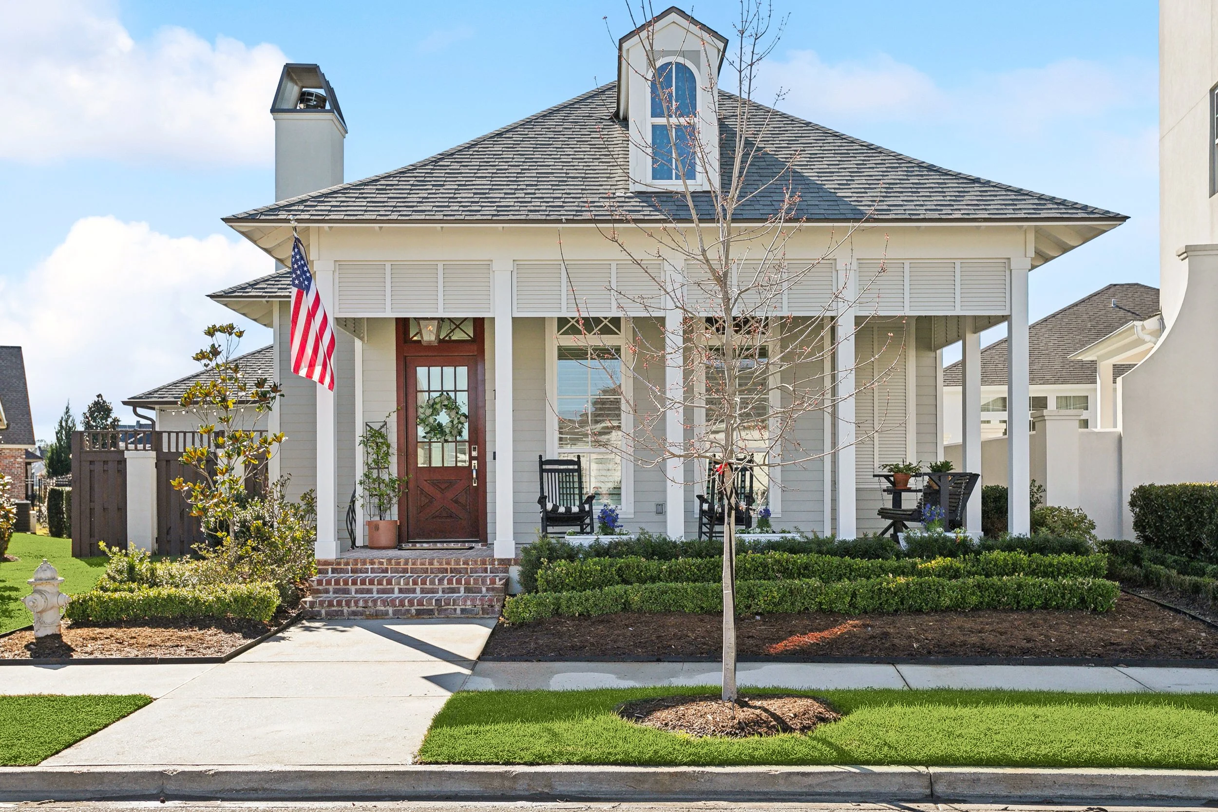 Front view of a white house with a brick porch, American flag, leafless tree, and green lawn.